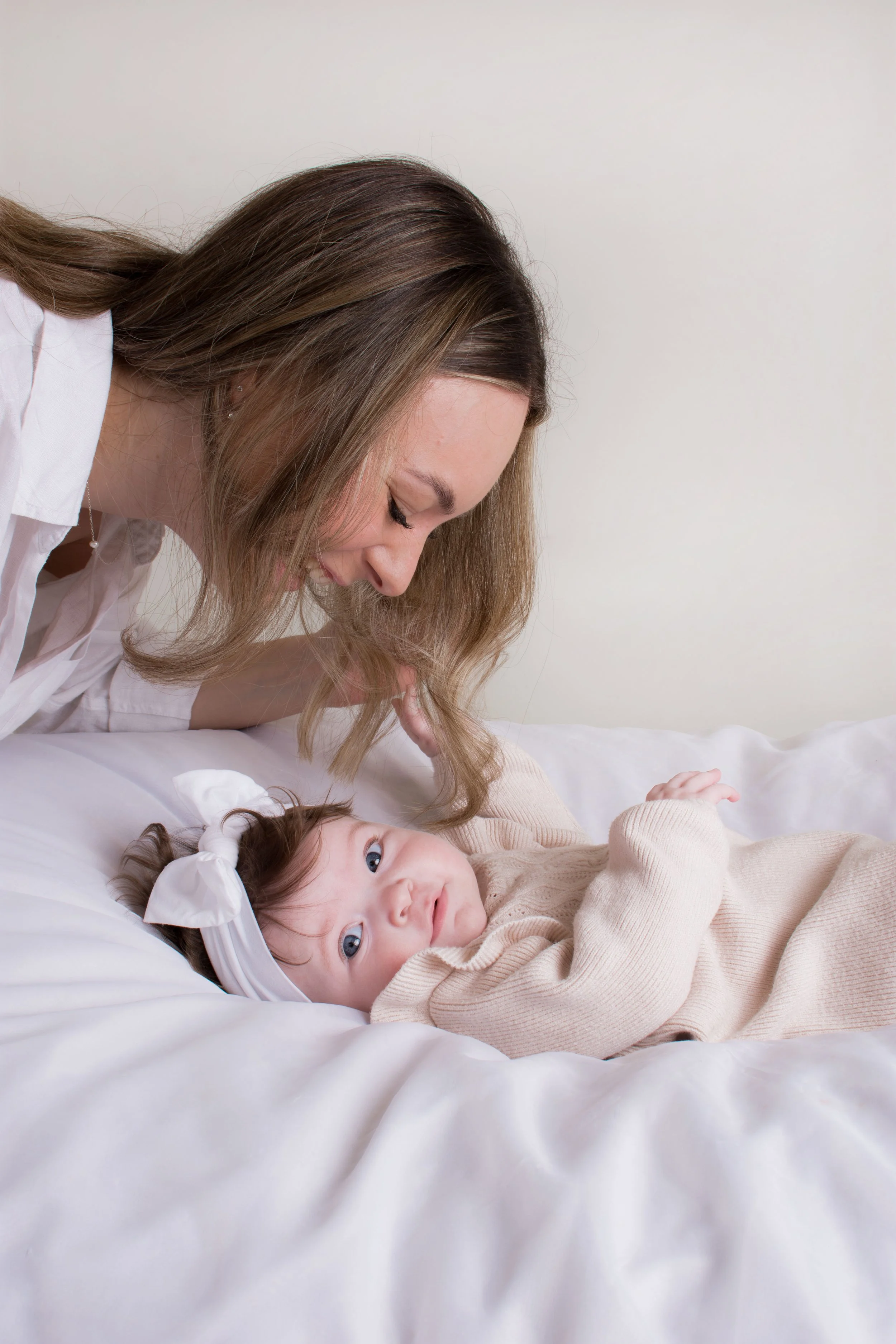 A woman leaning over a young girl lying on a bed, both looking at each other affectionately. The girl is wearing a beige sweater and has large blue eyes and dark curly hair with a white bow. The woman has long brown hair and is wearing a white shirt. The background is plain and light-colored.