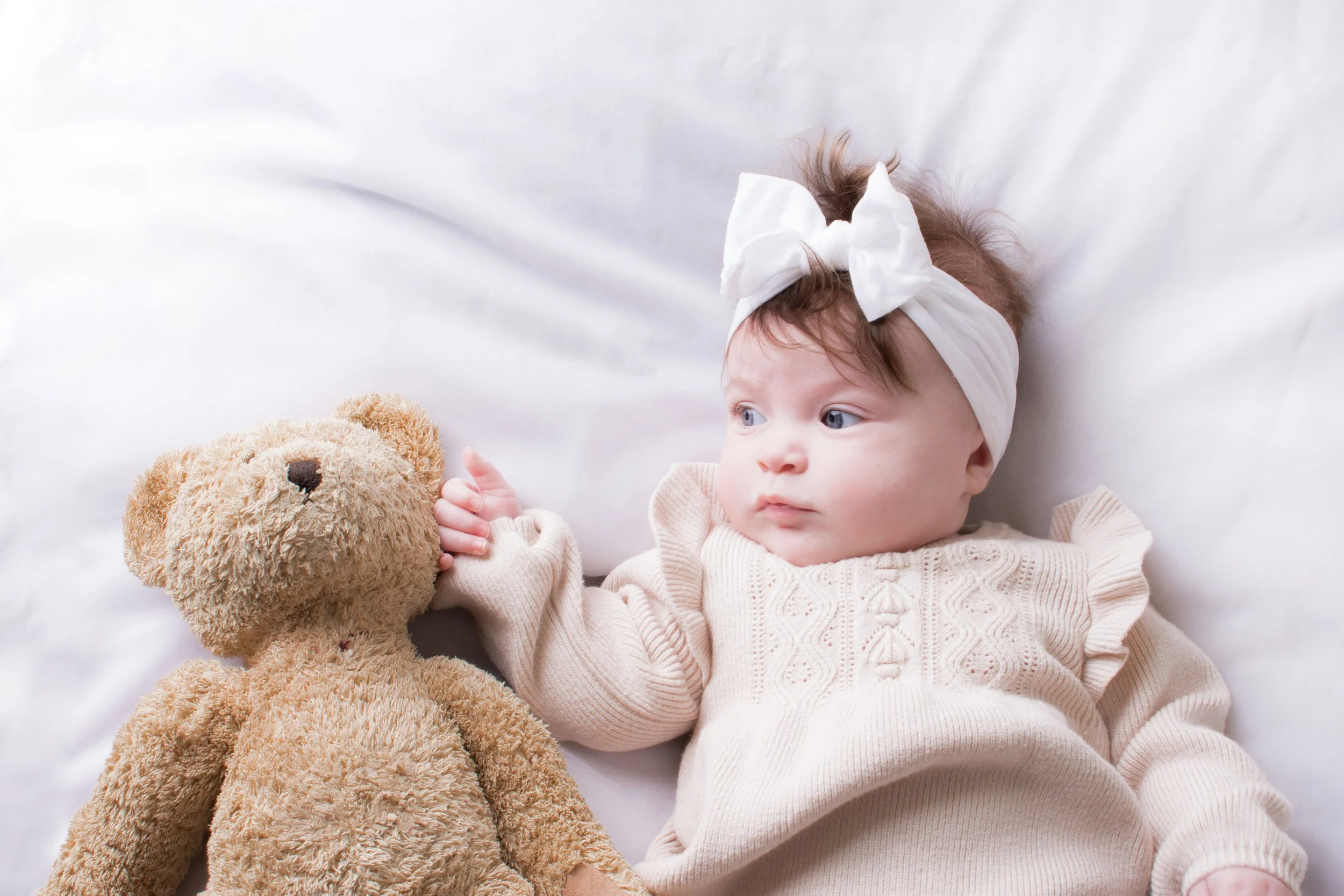A baby girl with light skin, blue eyes, and brown hair, lying on a white bed with a white headband with a large bow, holding a beige teddy bear.