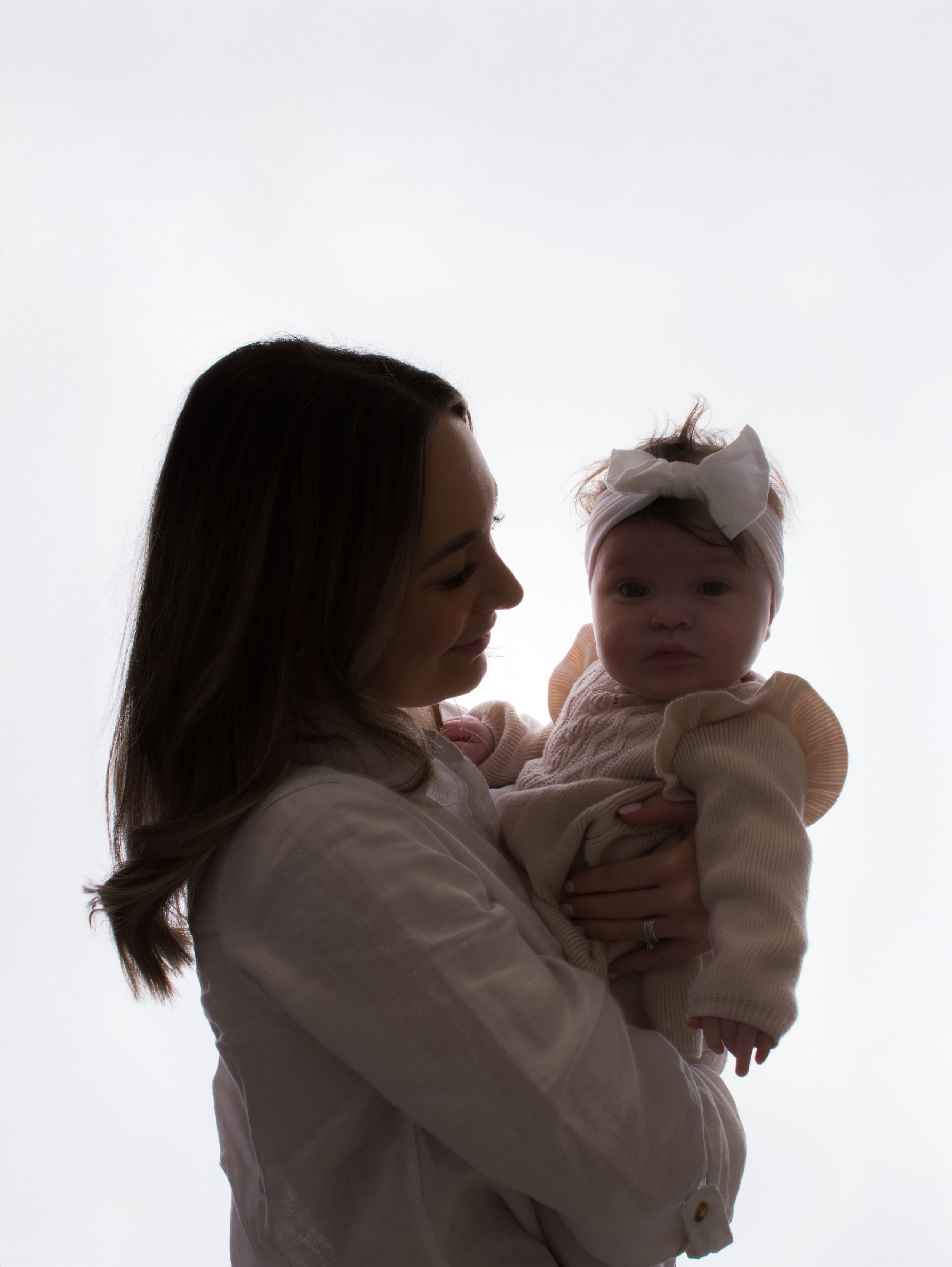 A woman with long dark hair holding a baby with a headband and light-colored sweater, both silhouetted against a light background.