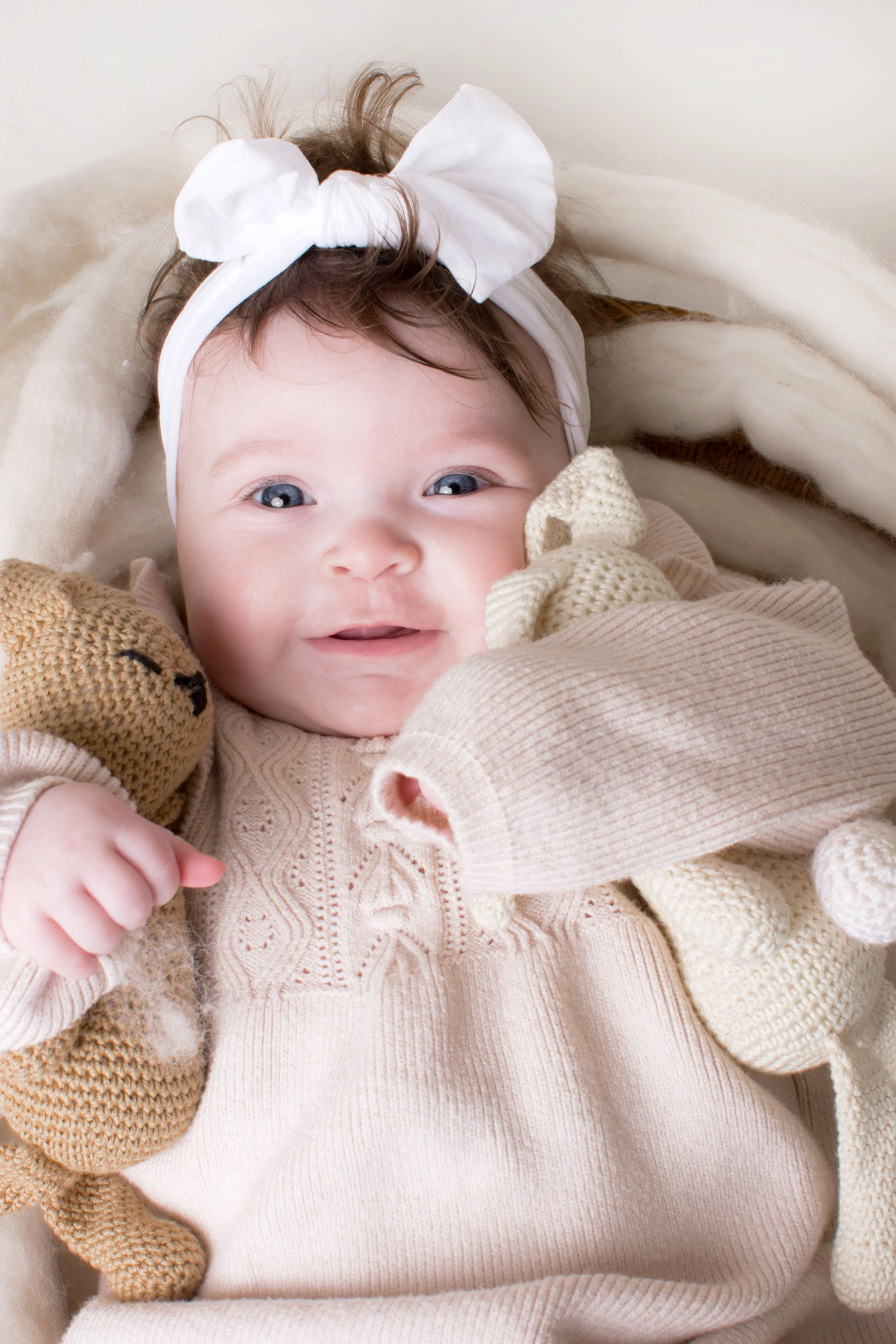 A smiling baby girl with blue eyes and brown hair, wearing a white headband with a bow, beige sweater, and holding a knitted giraffe plush toy, lying on a cream-colored blanket.
