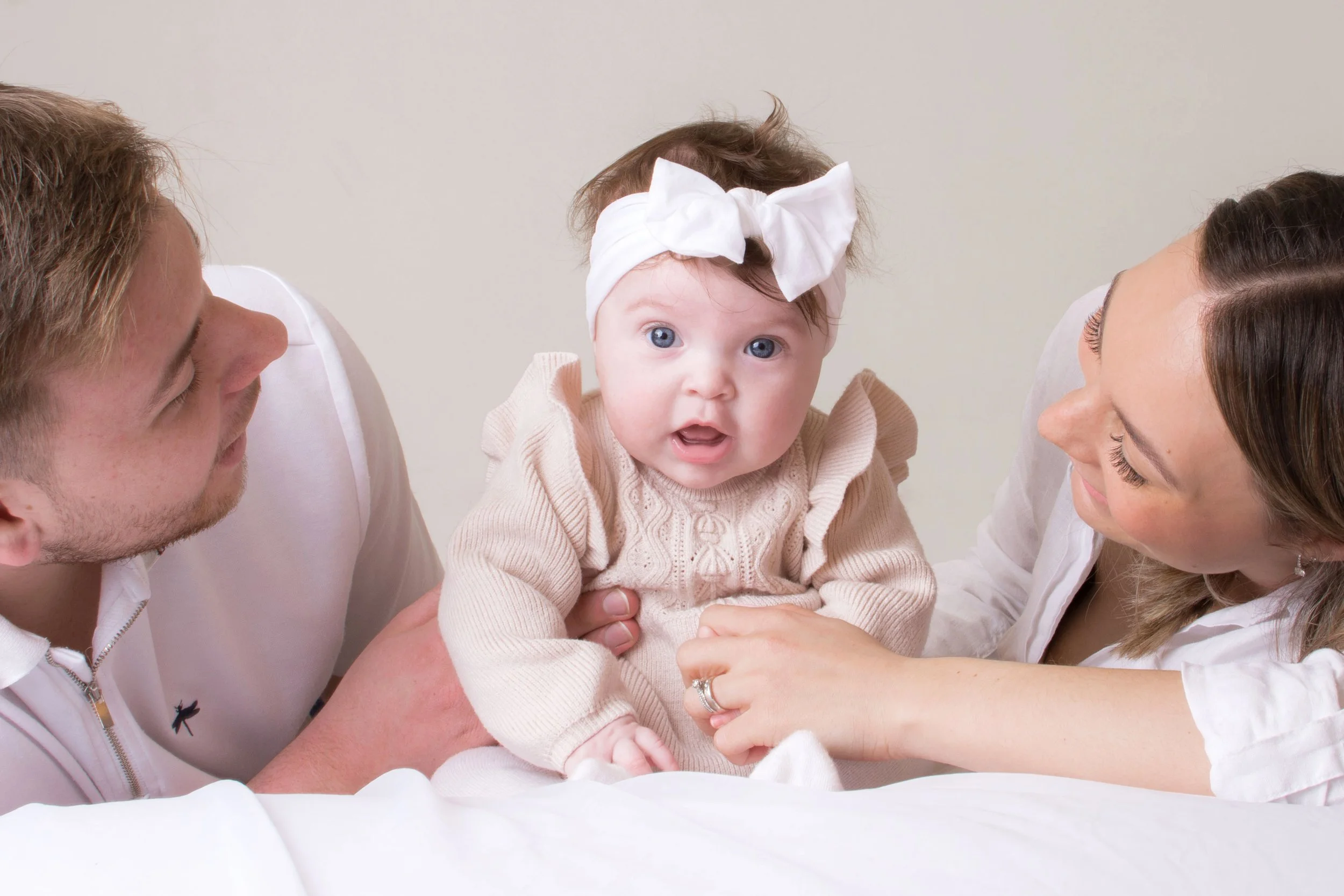 A baby girl with blue eyes, wearing a beige sweater and a white headband with a bow, is being held by an adult. She faces forward with an open mouth. Two adults, a man on the left and a woman on the right, are leaning in close to her, smiling. The background is plain and light-colored.