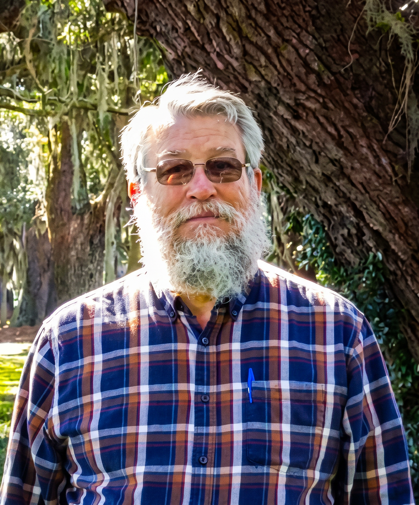 A man with gray hair, beard, and sunglasses standing outdoors in front of a large tree with green leaves, wearing a plaid shirt and holding a blue pen in his breast pocket.