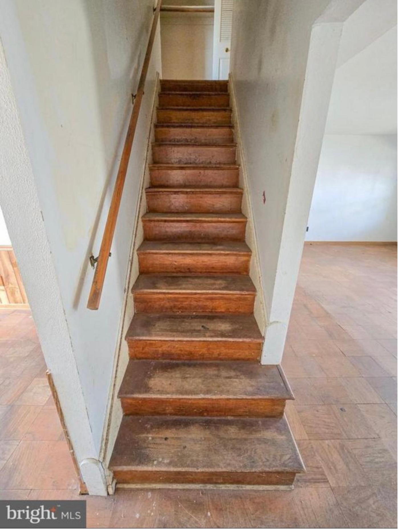 A wooden staircase with 12 steps and a wooden handrail on the left side, situated against white walls inside a house with hardwood flooring.