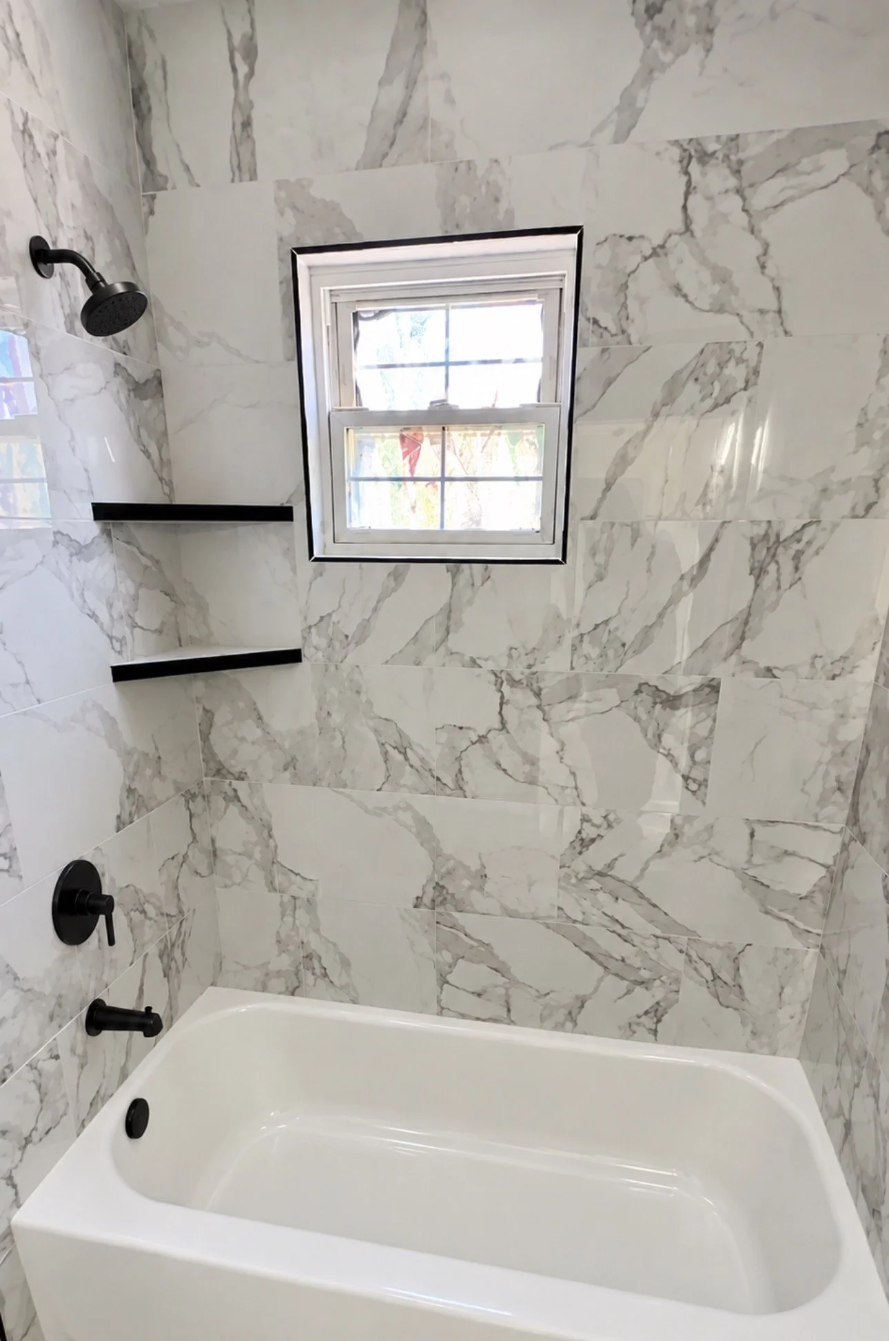 Bathroom shower area with marble tile walls, a black showerhead and handles, a small window with a grid, and a white bathtub at the bottom.