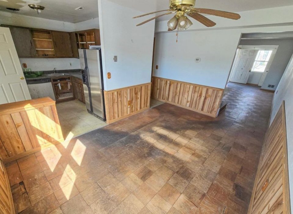 Empty room with wood paneling halfway up the walls, a ceiling fan, and hardwood flooring with discoloration; an adjacent kitchen visible through an open doorway.