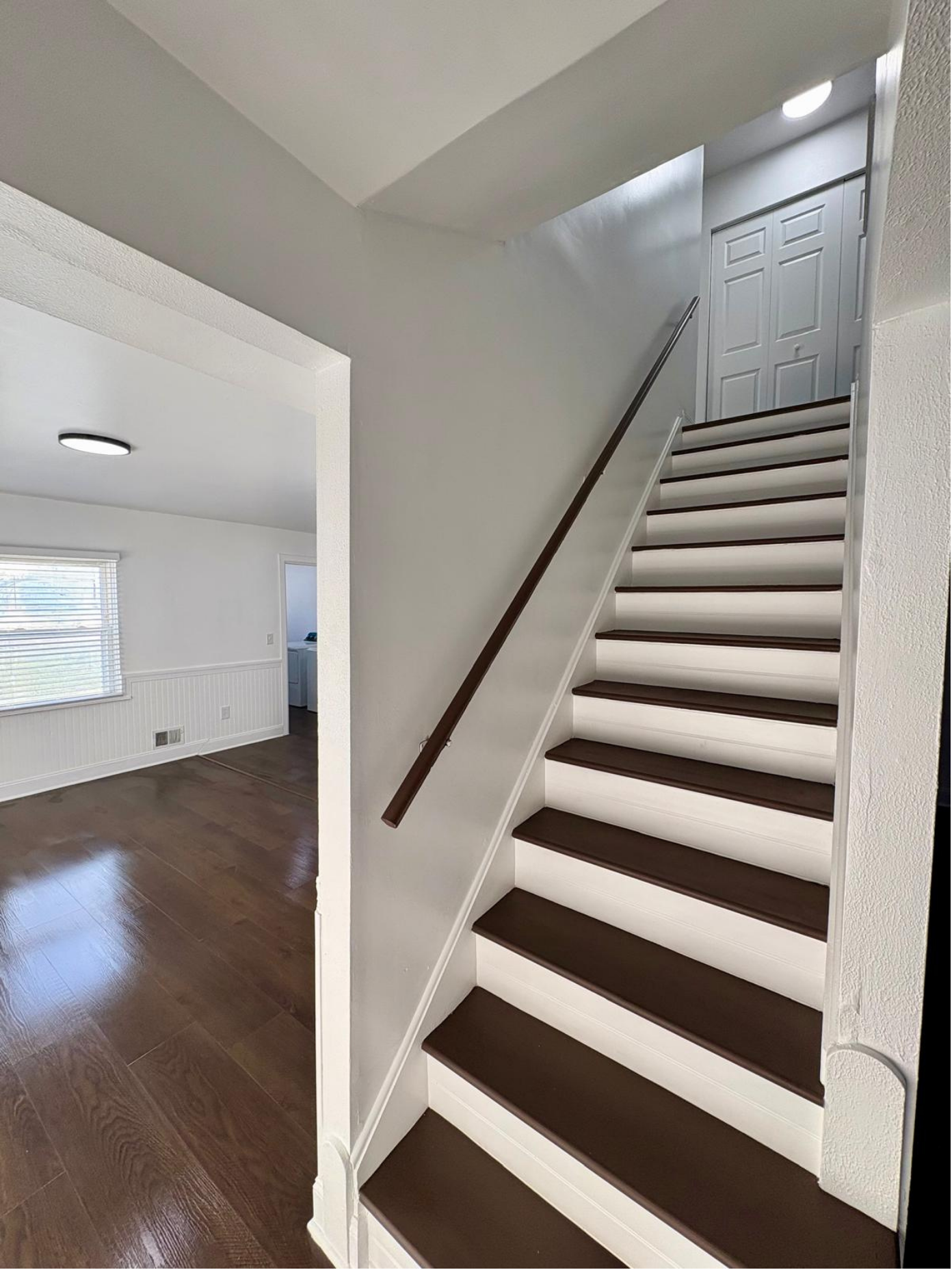 Interior view of a home showing a staircase with dark brown steps and white risers, a brown handrail, and a white wall. Adjacent room has wood flooring, a window with blinds, and white walls.