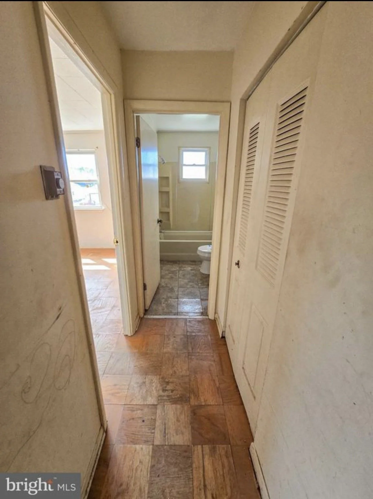 Hallway leading to a bathroom with a small window, and a room with a window and wooden flooring to the left.