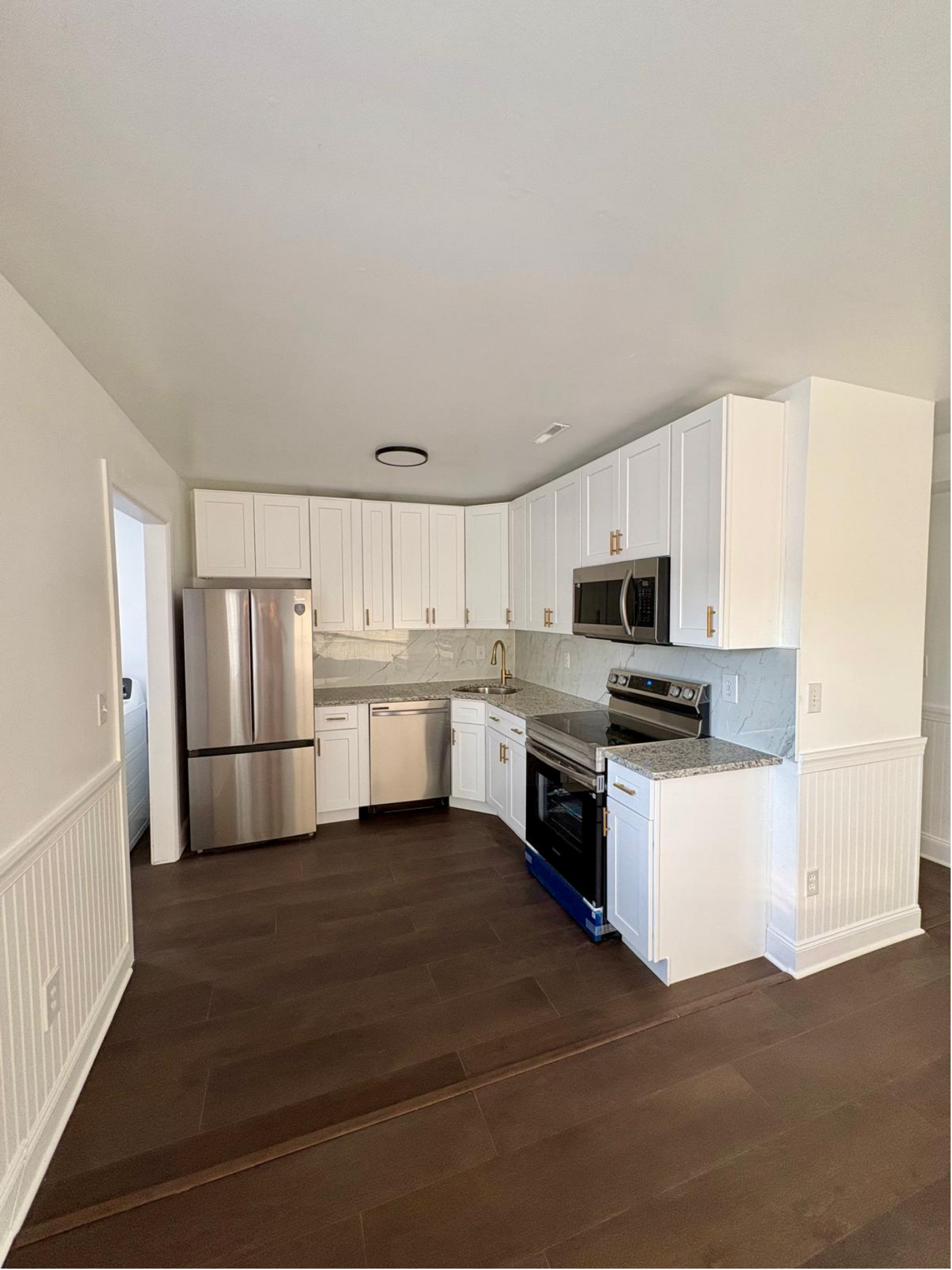 Modern kitchen with white cabinets, stainless steel appliances including a refrigerator, microwave, and oven, granite countertops, and dark wood flooring.