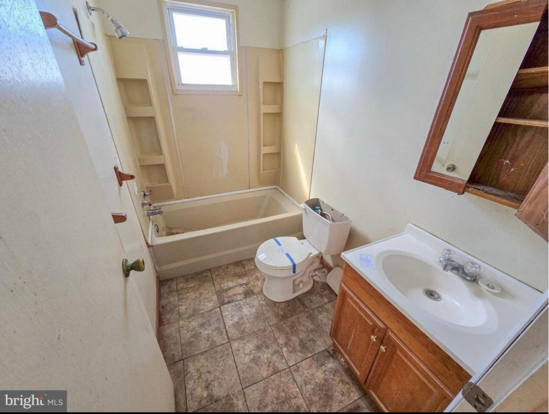 A bathroom with a vanity with a sink and wooden cabinet on the right, a toilet next to it with a toilet paper holder, a shower with a glass door, a window above the shower, and built-in shelves in the wall. The floor is tiled with brown and beige tiles.