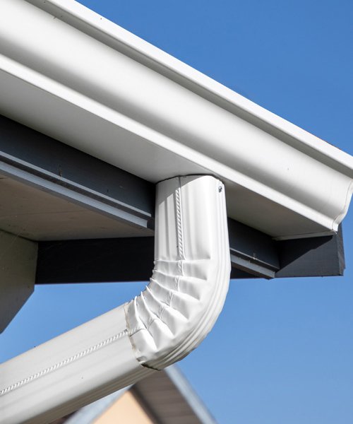 Close-up of a white gutter system with a curved pipe connecting to the downspout underneath a roof overhang, against a clear blue sky.