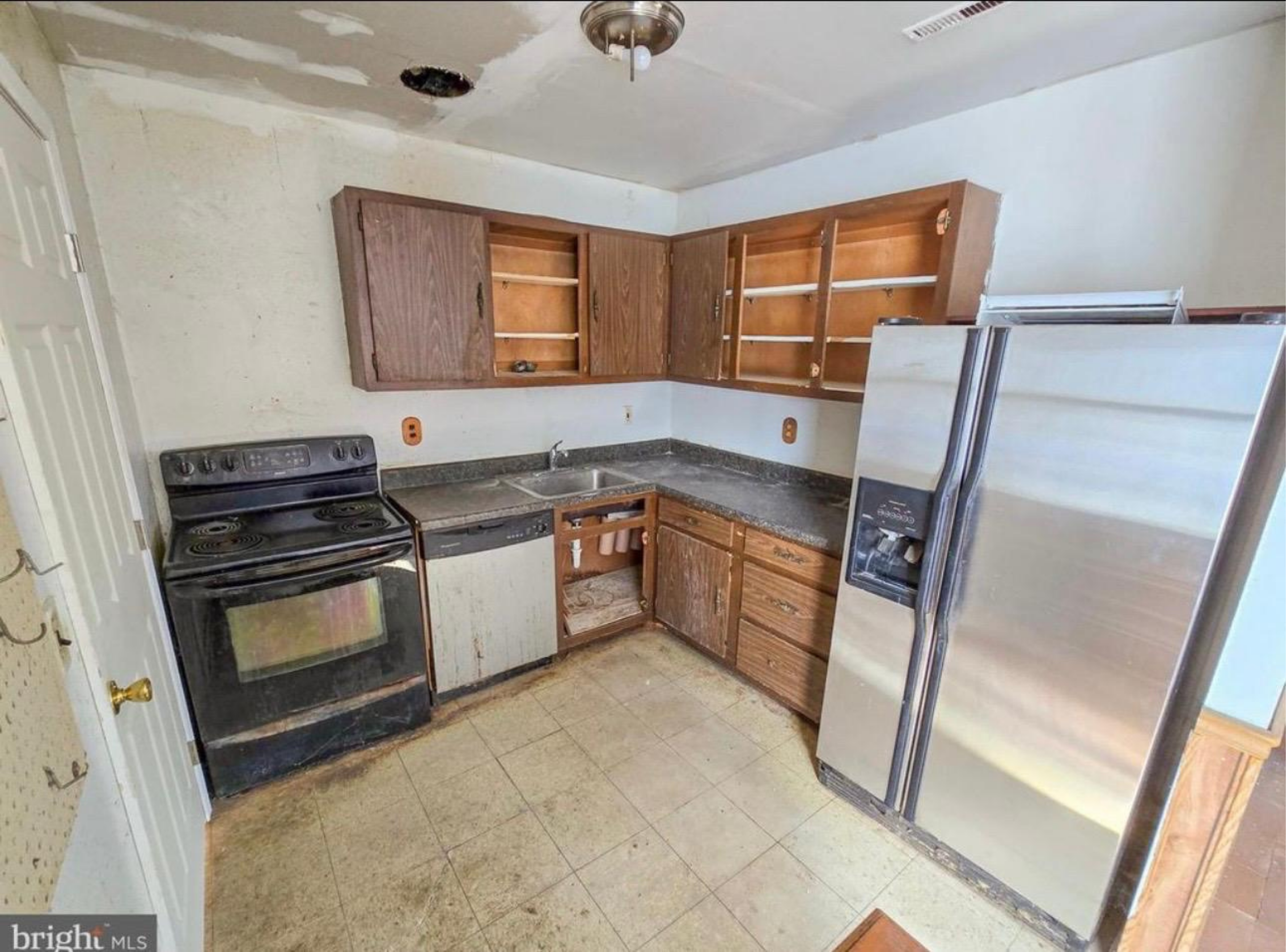 Empty kitchen with damaged upper cabinets, missing doors, and peeling ceiling paint. Black stove and a side-by-side refrigerator are visible.