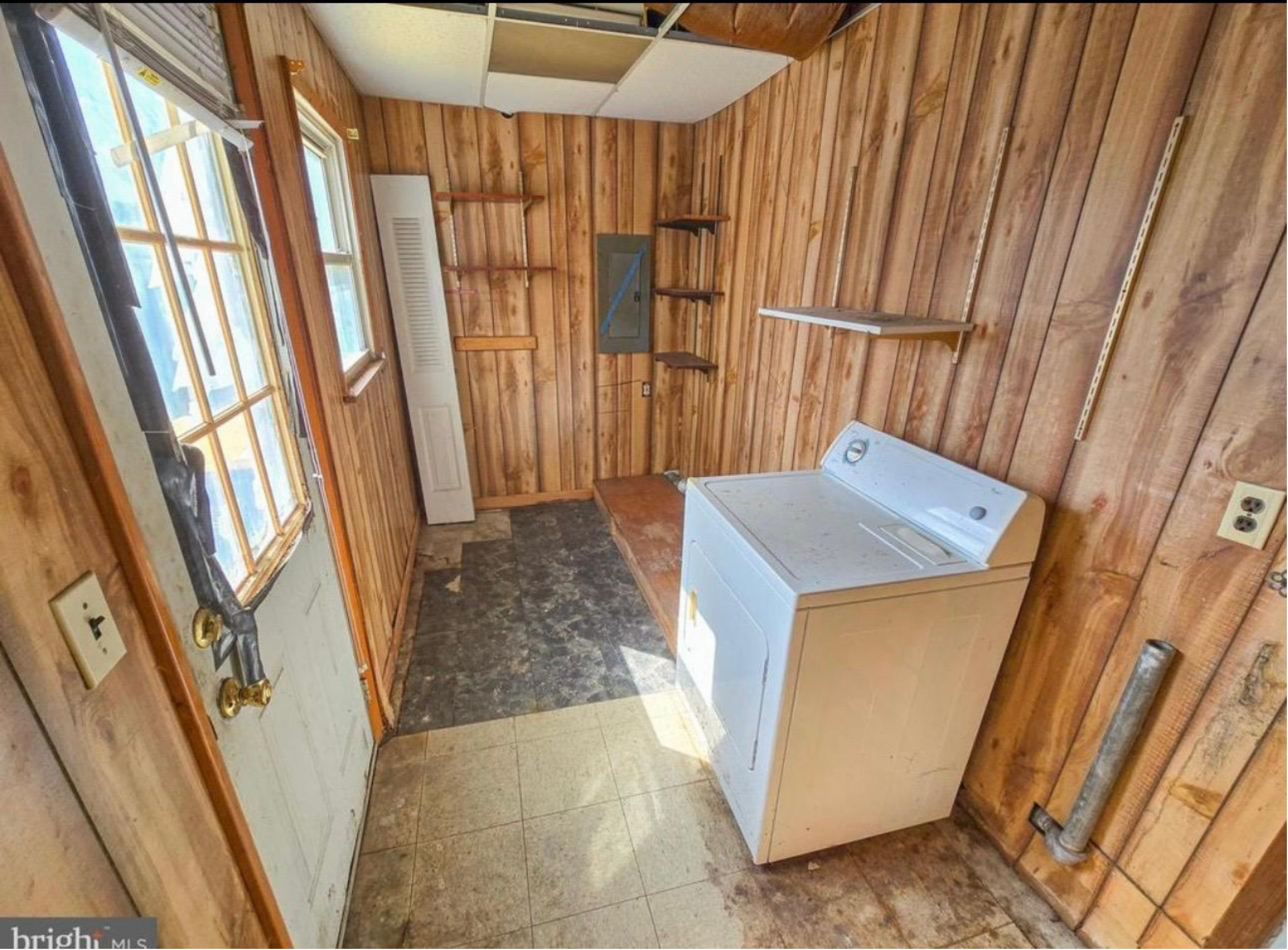 Interior of a woodworking or renovation space with wood-paneled walls, windows, and exposed pipes, showing an unfinished or in-progress room with shelves and construction materials.