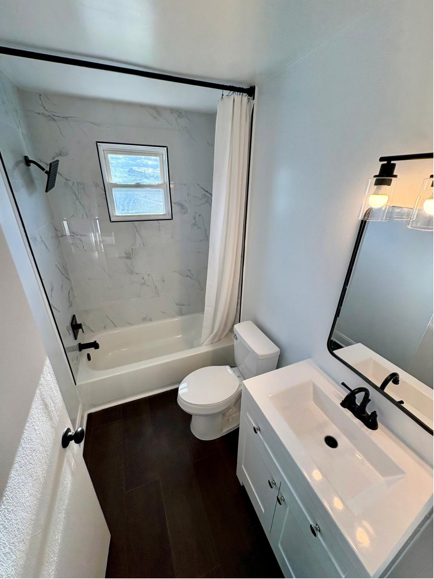 A modern bathroom with a bathtub and shower, a small window, a white toilet, a white vanity with a black faucet, a large mirror, and dark tiled flooring.