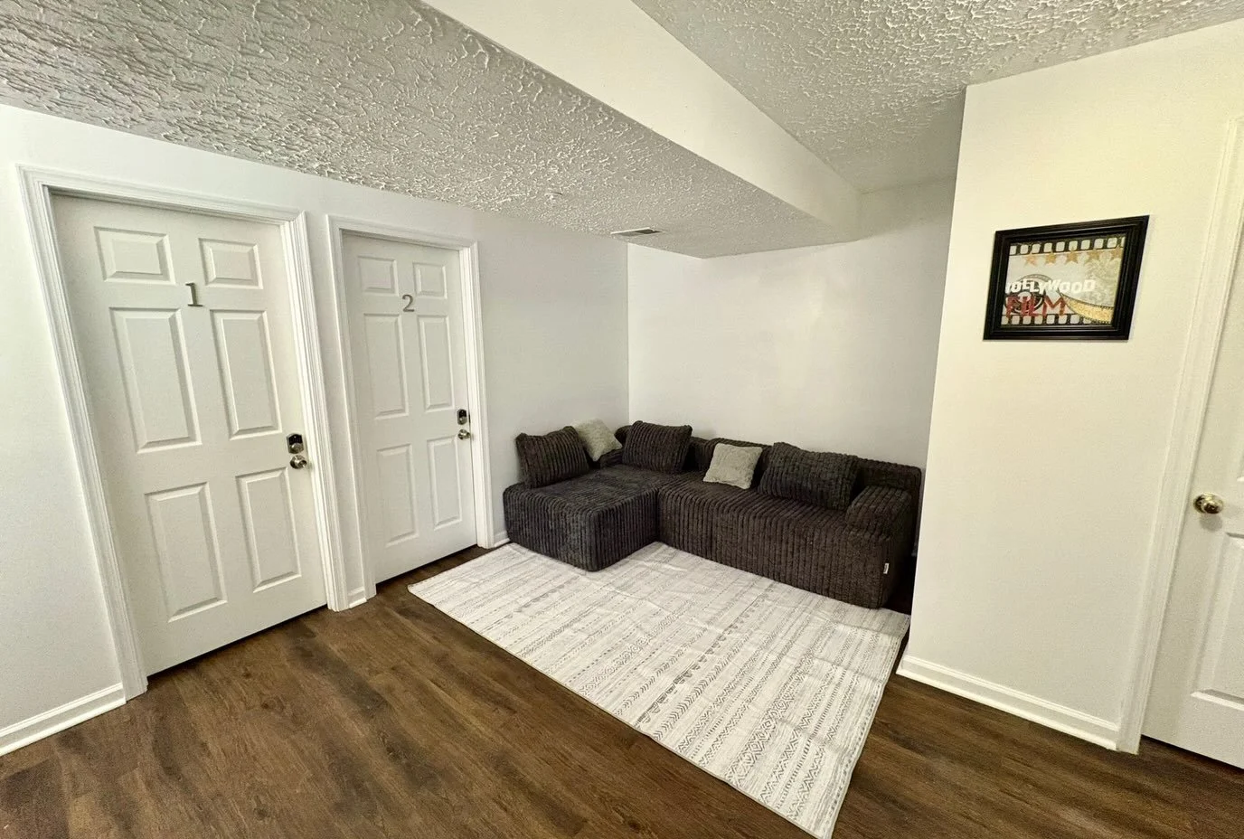 Living room with two closed white doors labeled 1 and 2, a dark brown sectional sofa with pillows, a white rug, and a small framed picture on the wall.
