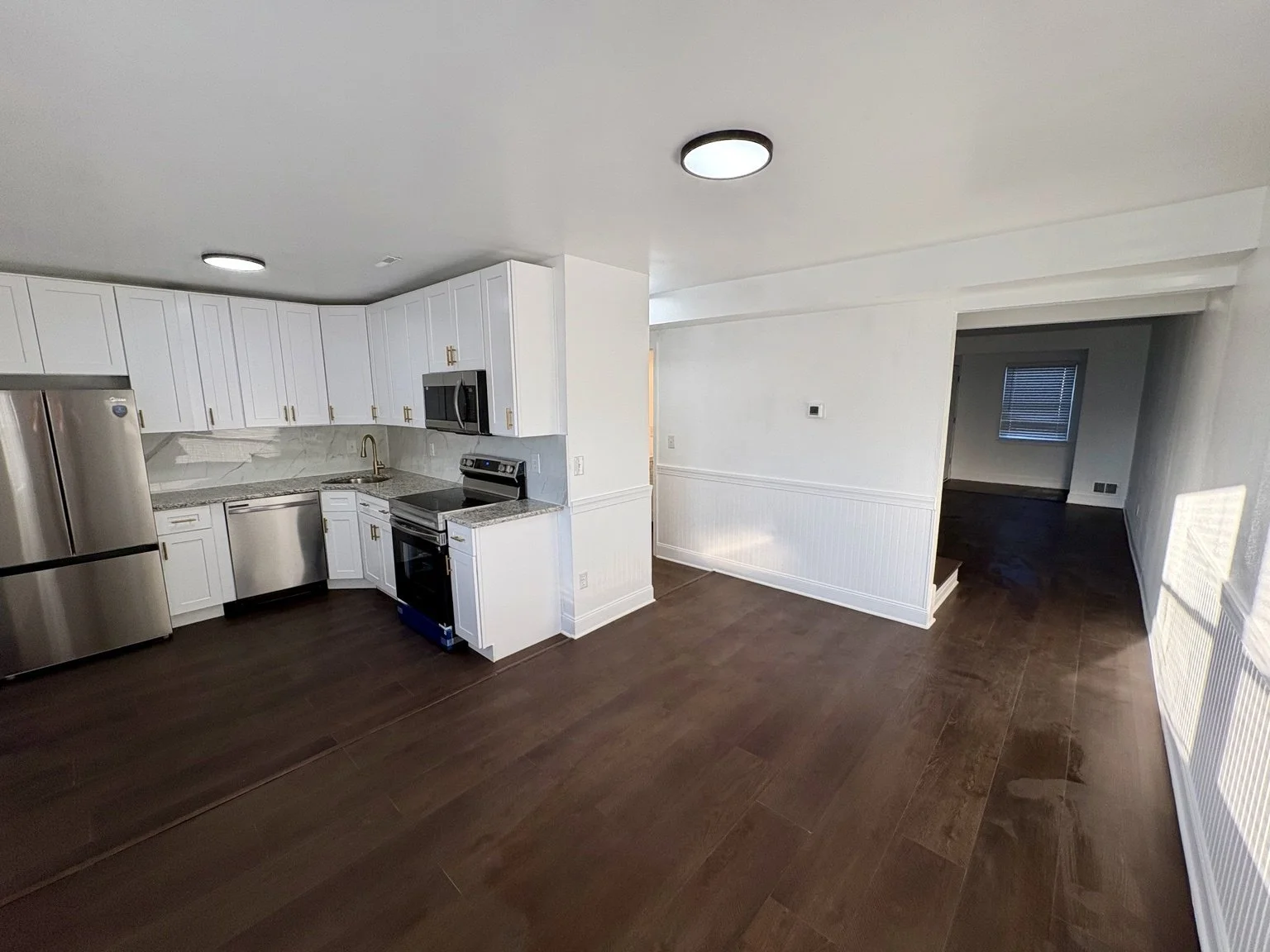 Modern kitchen with white cabinets, stainless steel appliances, dark hardwood flooring, and an adjacent empty room with a small window.
