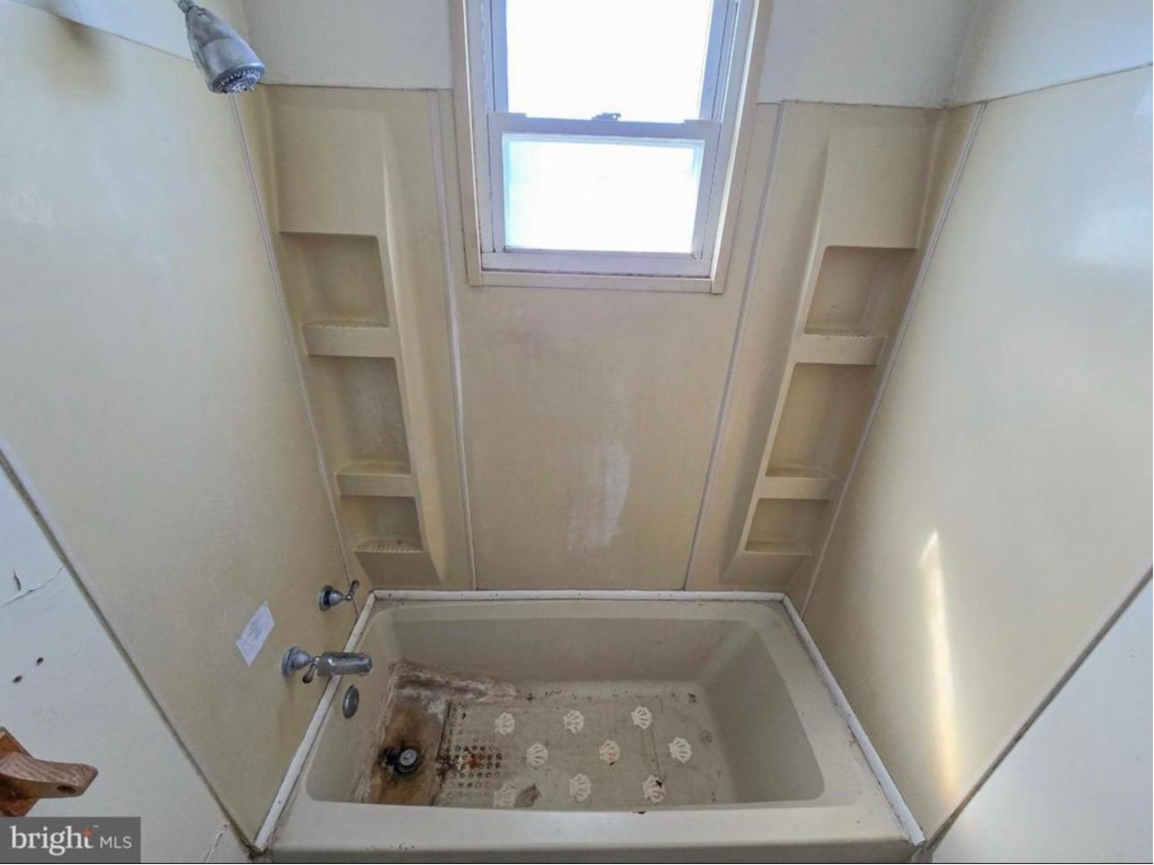 A bathroom with a built-in corner tub, a window above it, and a shower with no door. The room appears unfinished, with visible water damage or dirt on the tub and walls.
