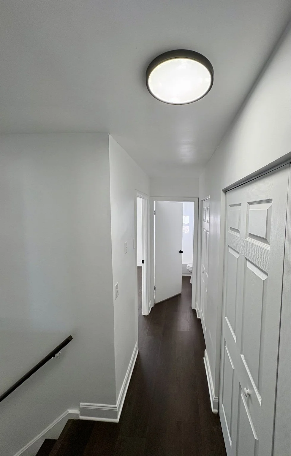 Bright hallway in a house with white walls, dark hardwood floors, and a ceiling light fixture, leading to a bathroom with an open door.