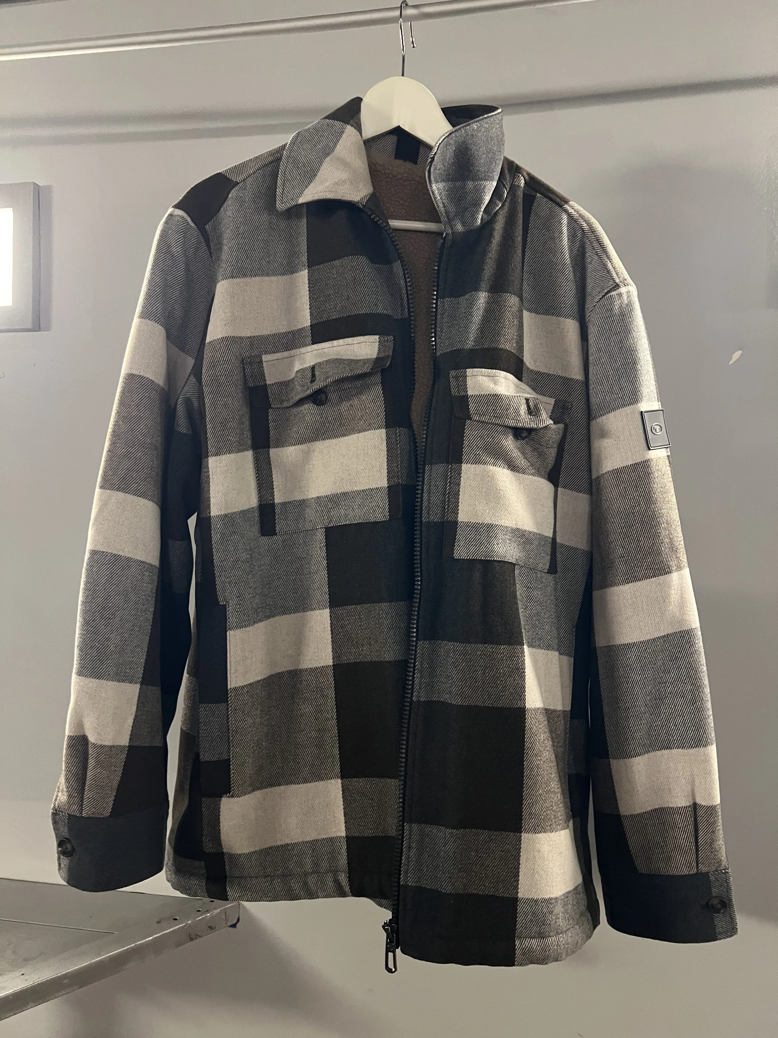Graues und schwarzes Karomuster Jacke mit Reißverschluss, Brusttaschen, Stehkragen und schwarzen Ärmelsäumen, hängt an einem Kleiderbügel.
