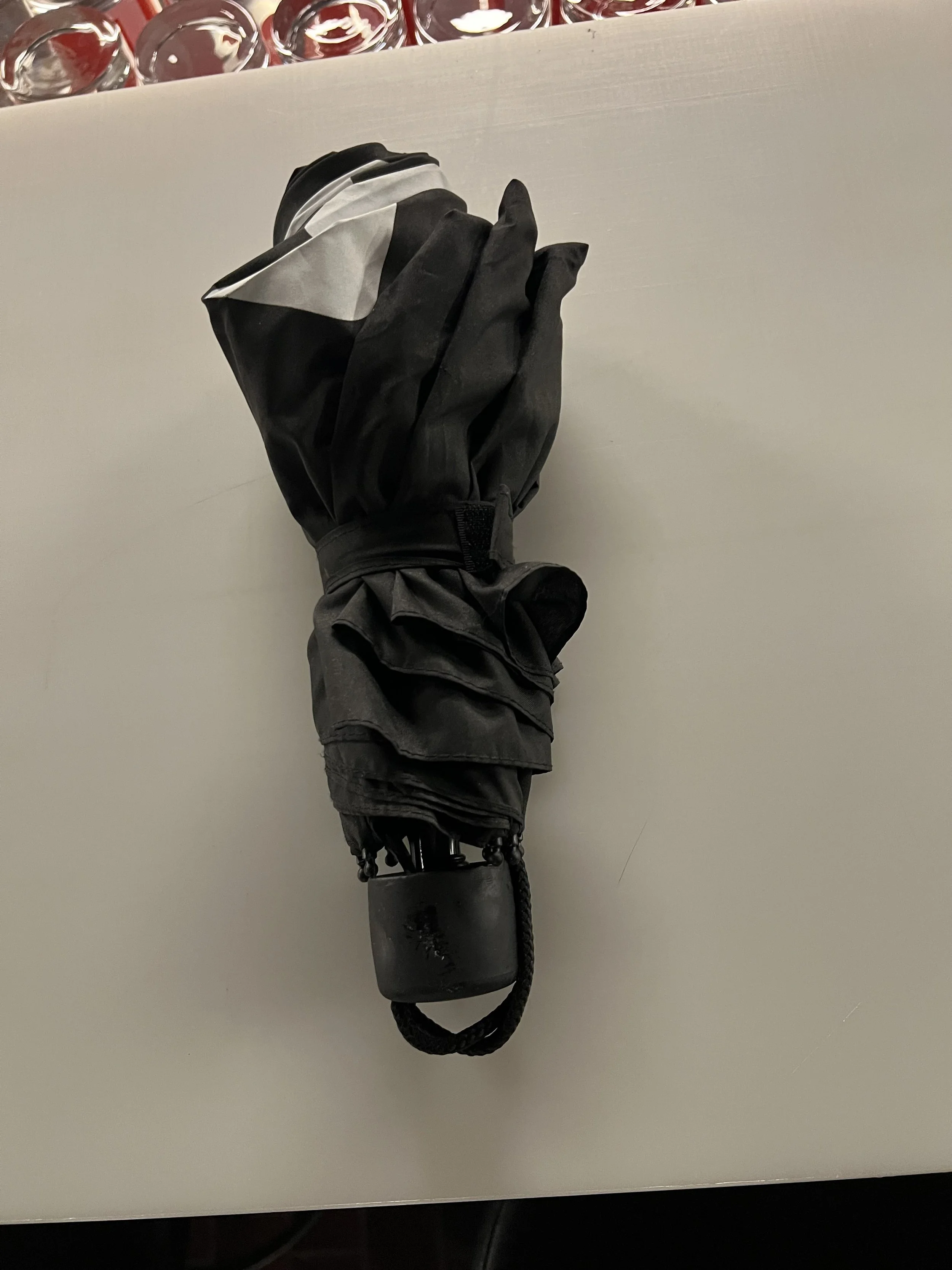 Ein schwarzer Regenschirm, eingepackt in eine schwarze und weiße Regenhülle, liegt auf einem weißen Tisch.