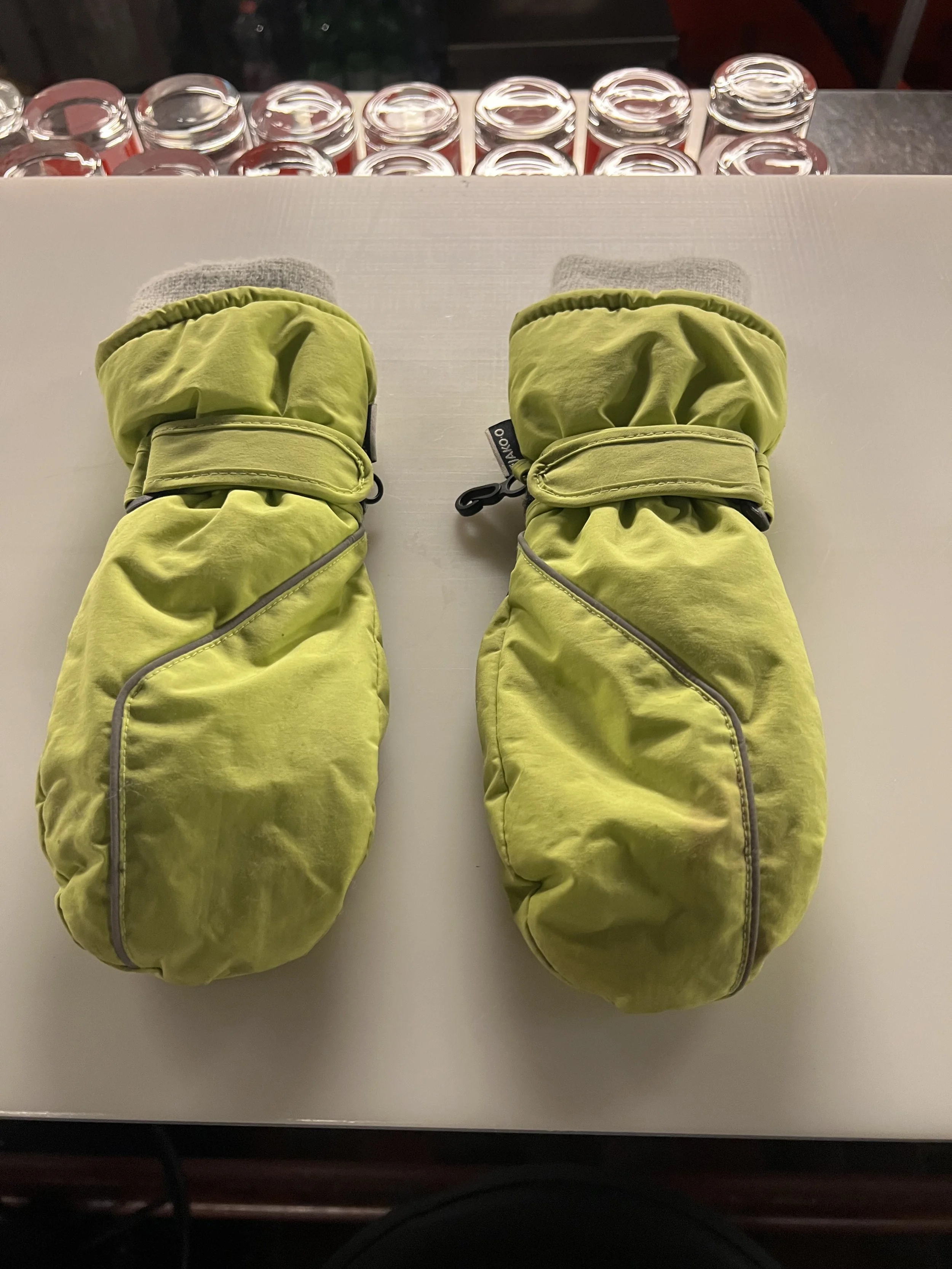 Zwei grüne Babyski-Handschuhe auf einem weißen Oberflächen, im Hintergrund sind einige Gläser.
