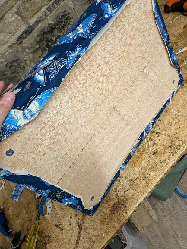 A person is sewing a piece of light-colored fabric with blue butterfly prints around a wooden frame on a workbench. The fabric is partially lifted, and sewing tools are visible nearby.