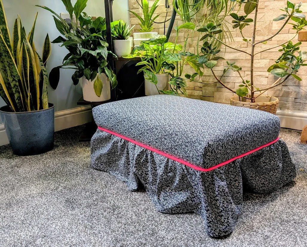 A cozy indoor nook with a black and white floral-patterned ottoman, surrounded by various potted green houseplants, and a textured beige brick wall in the background.