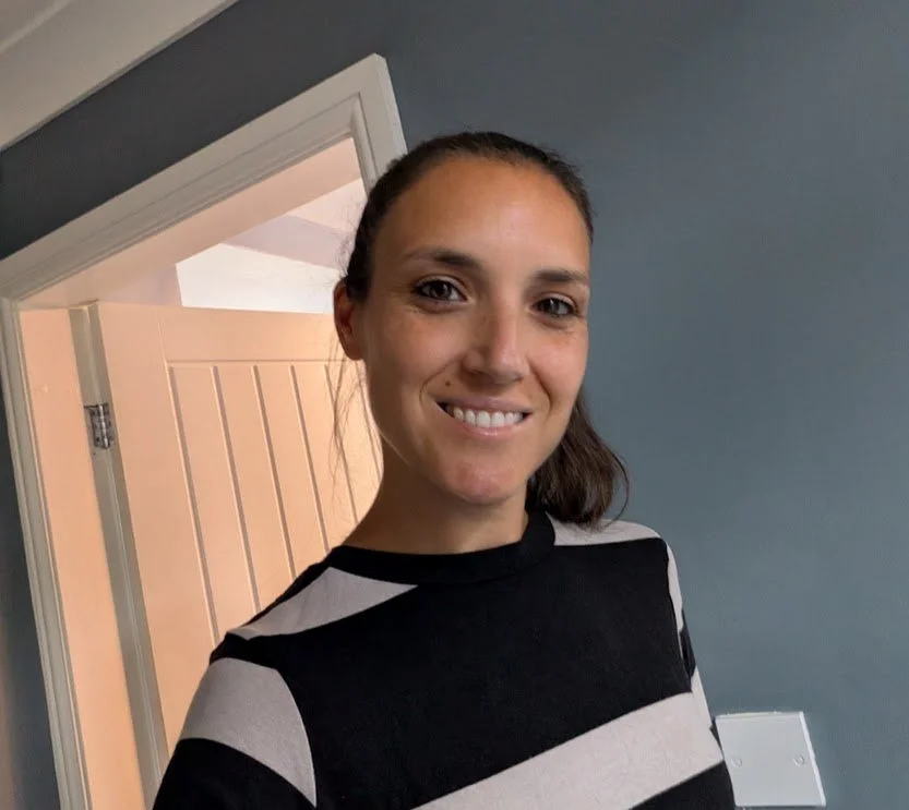 A young woman with brown hair in a ponytail, smiling, wearing a black and white striped shirt, standing inside a room with a blue wall and a door open to another room.