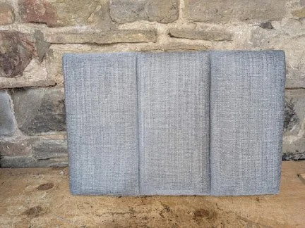 A light gray fabric upholstered headboard leaning against a stone wall on a wooden surface.
