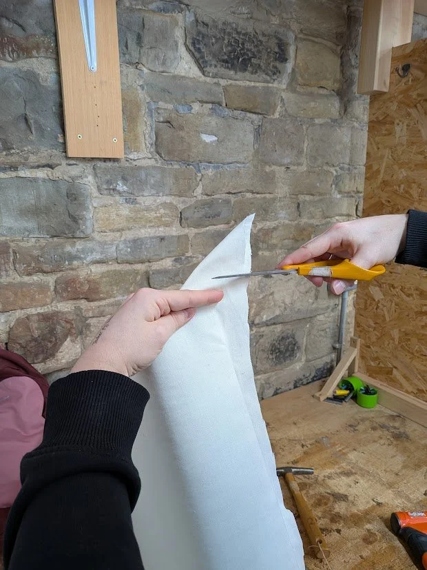 Someone cutting a piece of fabric with scissors while holding it with their other hand, in a workshop with a brick wall and wooden shelves.