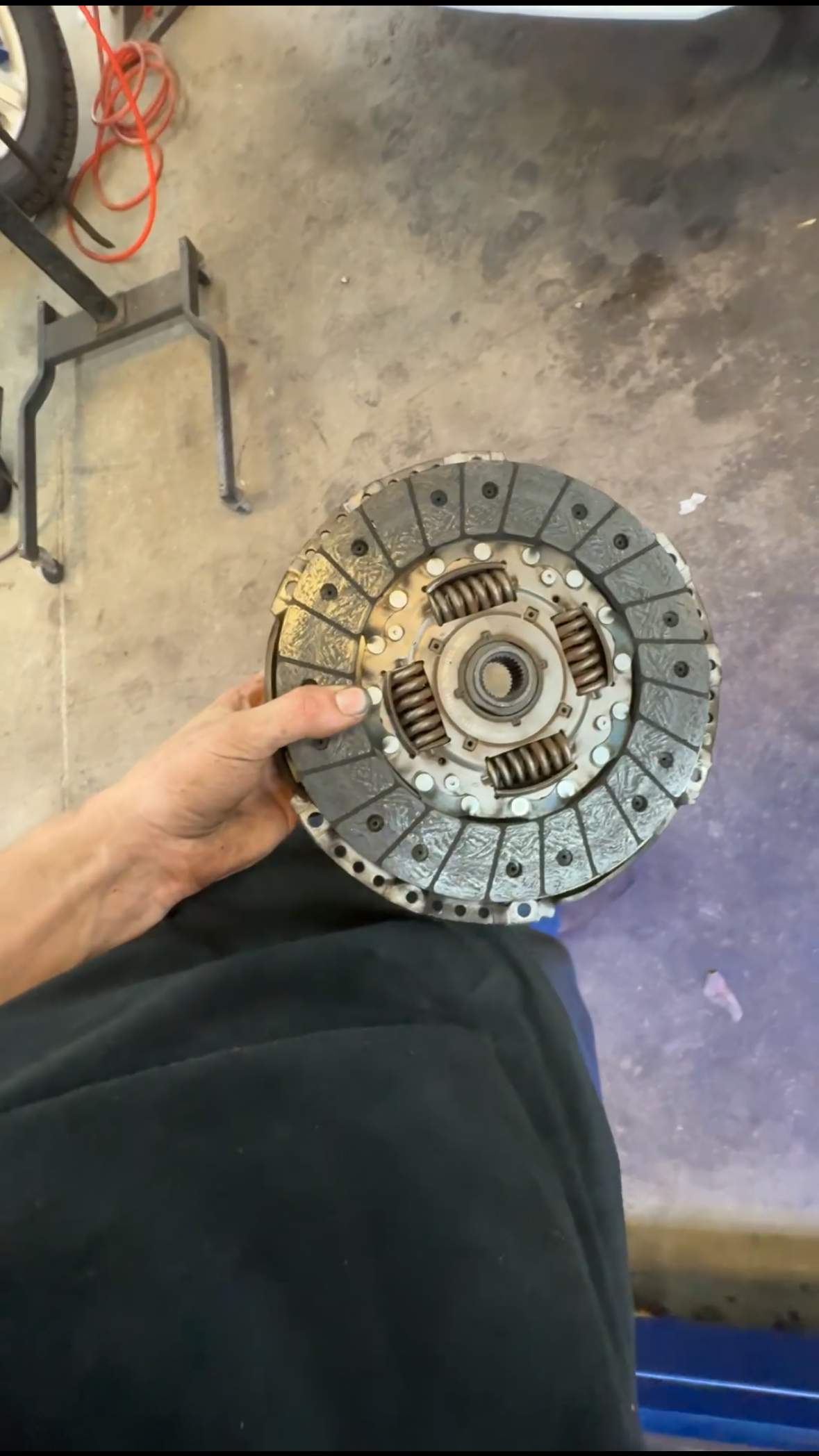 Hand holding a clutch friction disc, part of a vehicle's clutch assembly, in a garage or workshop setting.