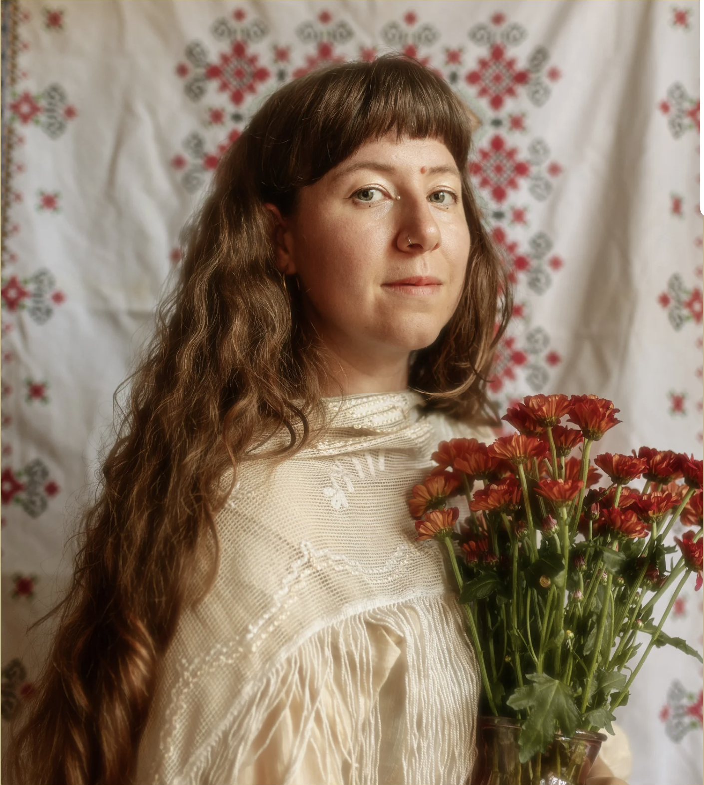 Foto von Luisa Melzig, einer Frau mit langen braunen Haaren. Sie sitzt vor einem rot bestickten Textil, in der Hand hat sie einen Blumenstrauß mit roten Chrysanthemen.