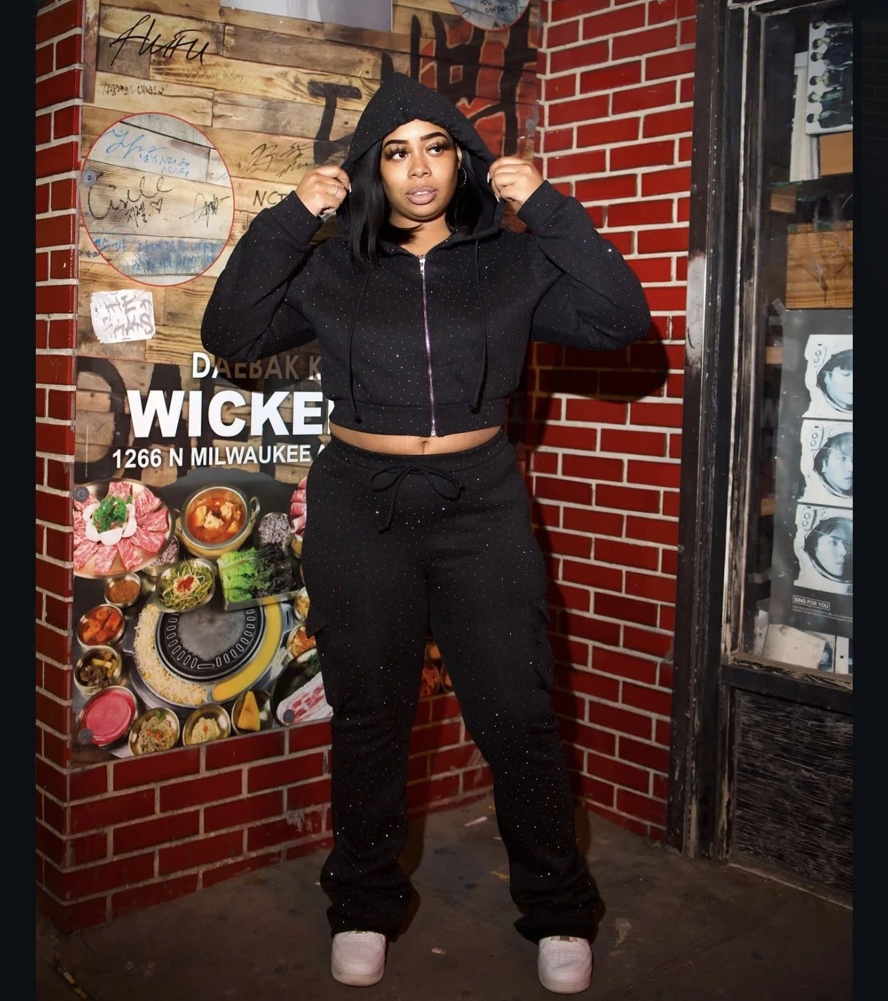 A woman wearing a black cropped hoodie with small white polka dots and matching black pants with small white polka dots, standing in front of a brick wall and restaurant sign, adjusting her hood with her hands.