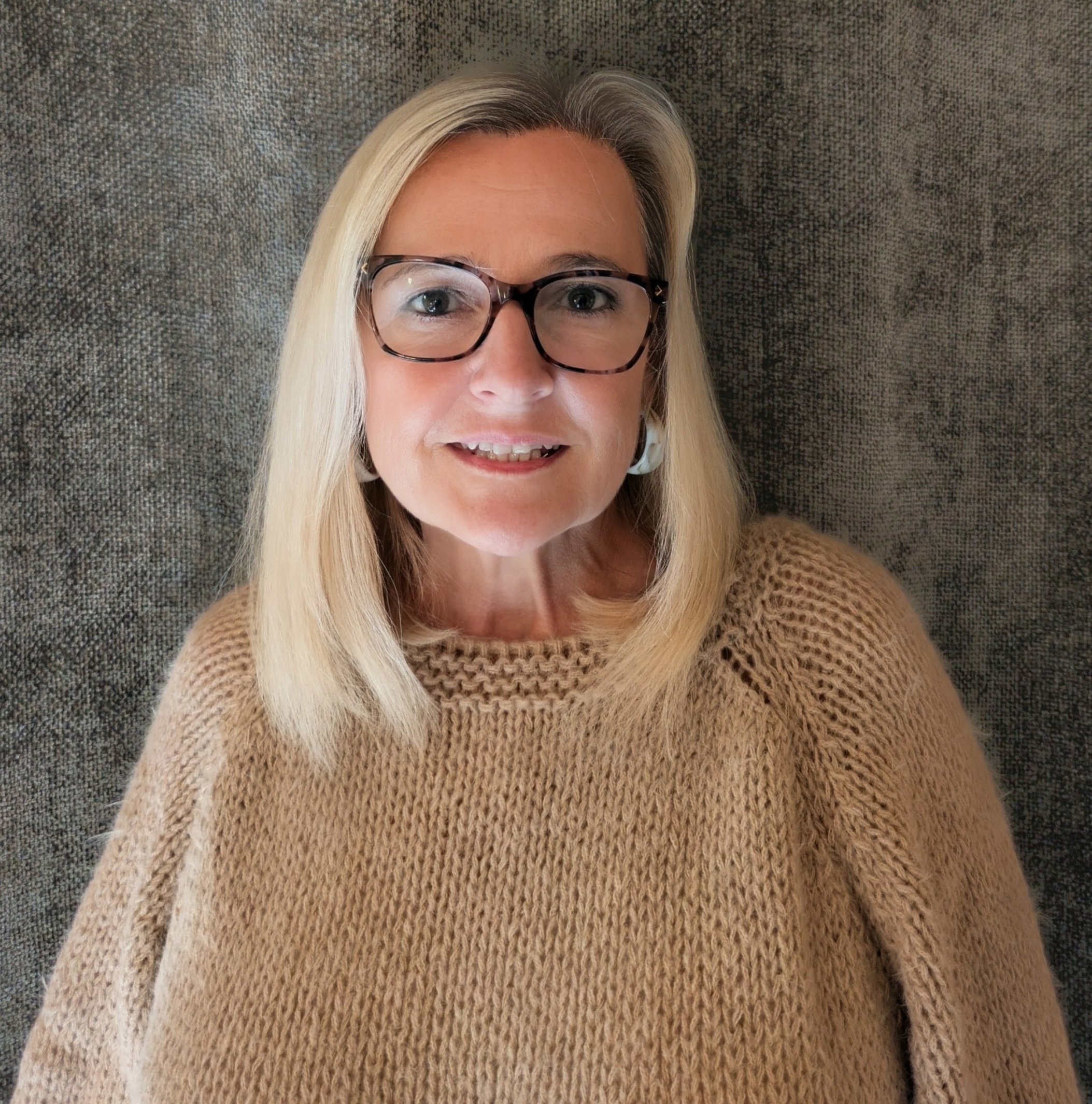 Smiling blonde woman wearing glasses and a tan knitted sweater in a close-up portrait