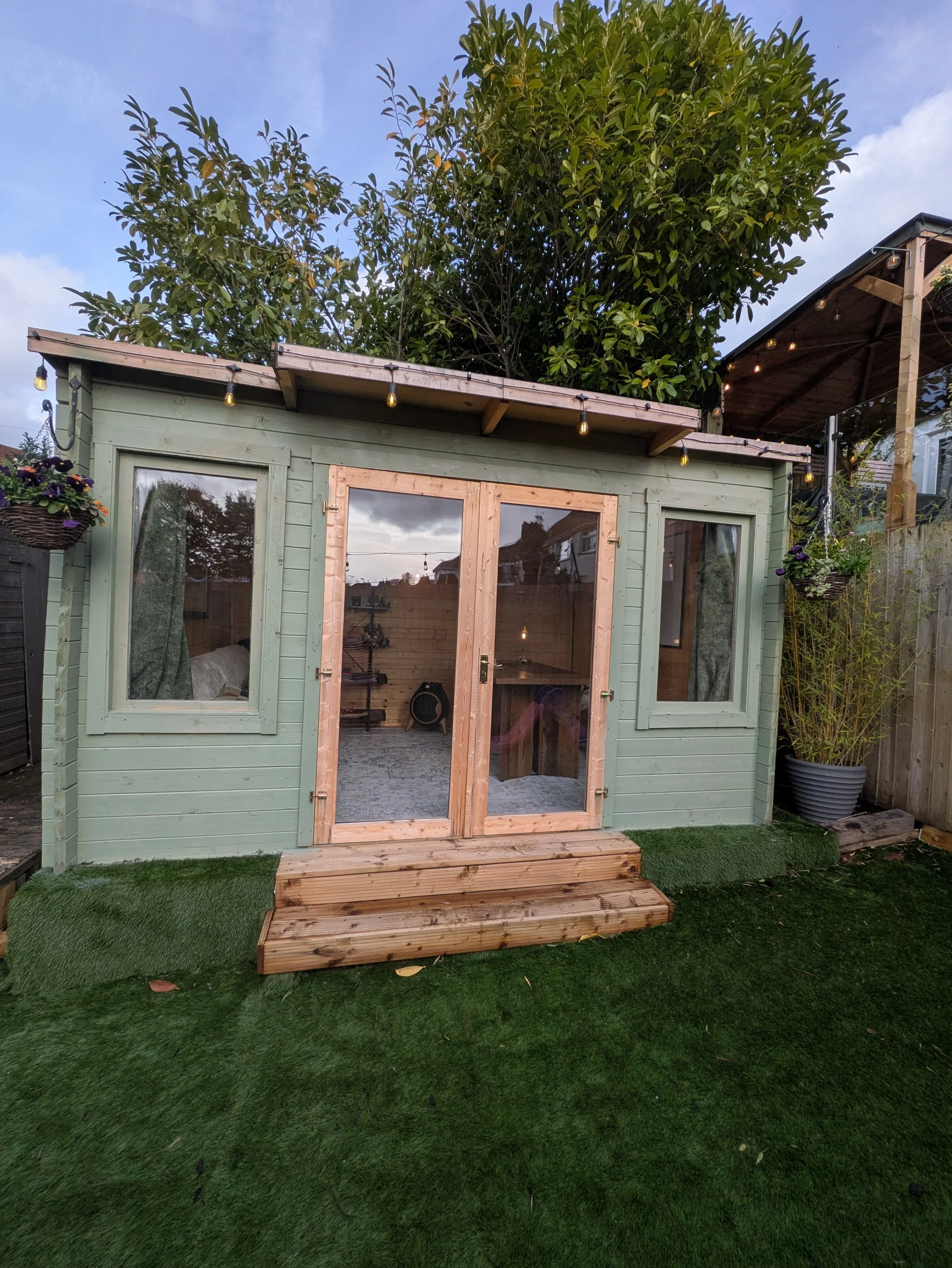 Green backyard garden room with glass double doors, wooden steps, string lights, potted plants, and surrounding trees
