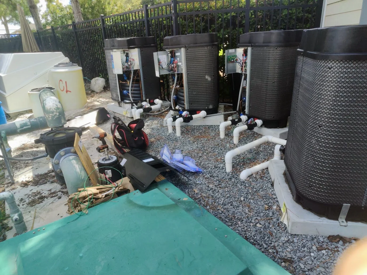 An outdoor Commercial Pool  setup with four large, black, metal-cased Heat pump pool heating units connected to white PVC piping, along with heavy duty commercial pool pumps and equipment.