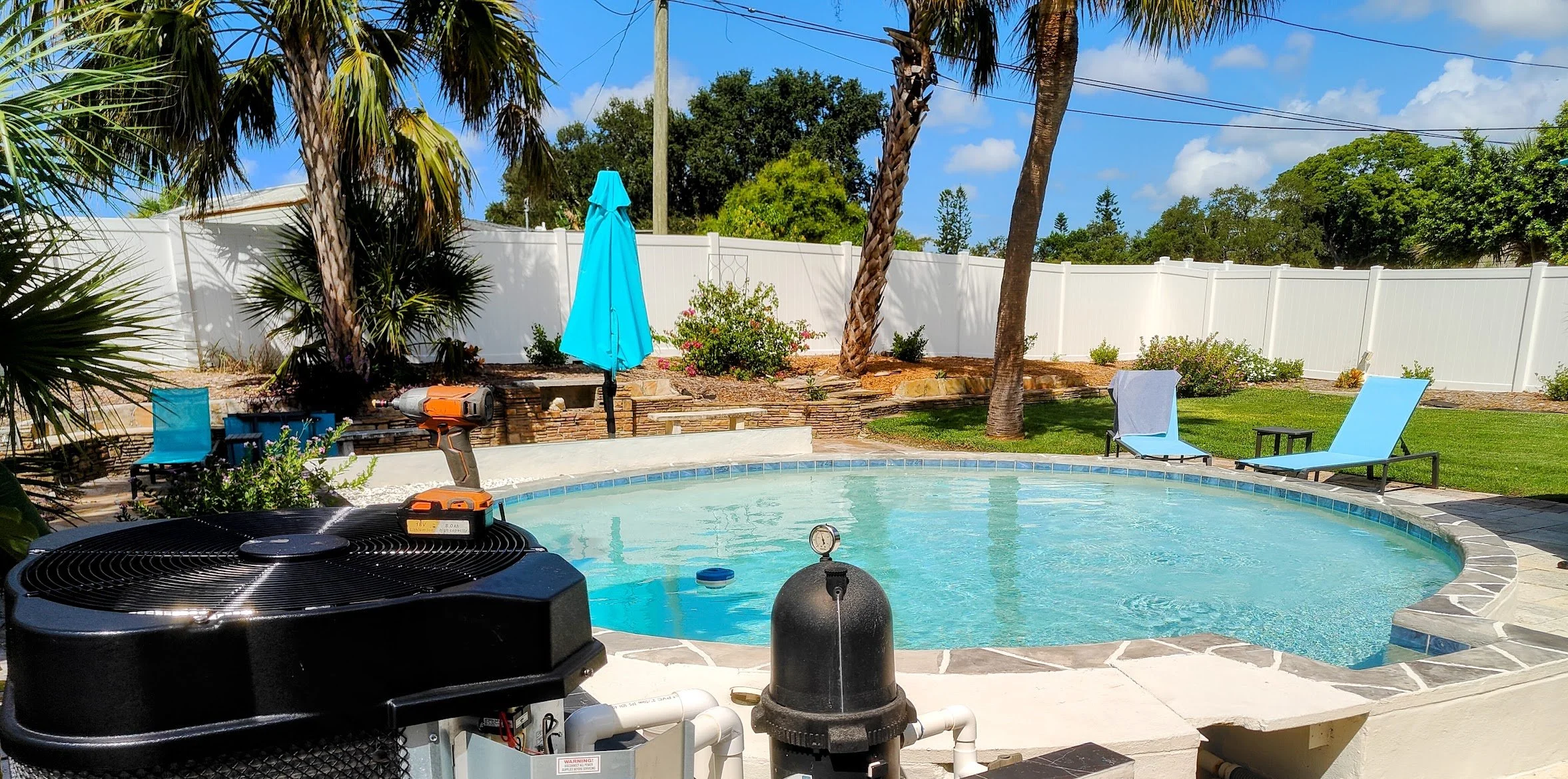 A backyard scene with a swimming pool, surrounded by a white fence, palm trees, and green shrubs. There are lounge chairs and a blue patio umbrella, with some pool equipment and a robot cleaner visible.