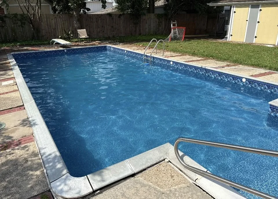 A backyard swimming pool with clear blue water, surrounded by a concrete deck, with a ladder at one end and a small hockey net at the far end, enclosed by a wooden fence and some trees.