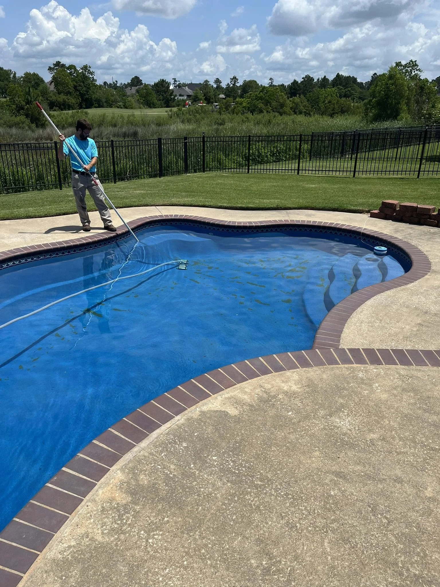 A man with a beard in a blue shirt and beige pants is cleaning a swimming pool with a long pole under partly cloudy skies. The pool is surrounded by a beige concrete deck with a dark brick border. There is a black metal fence in the background, with green grass, trees, and a golf course or large open area beyond it.