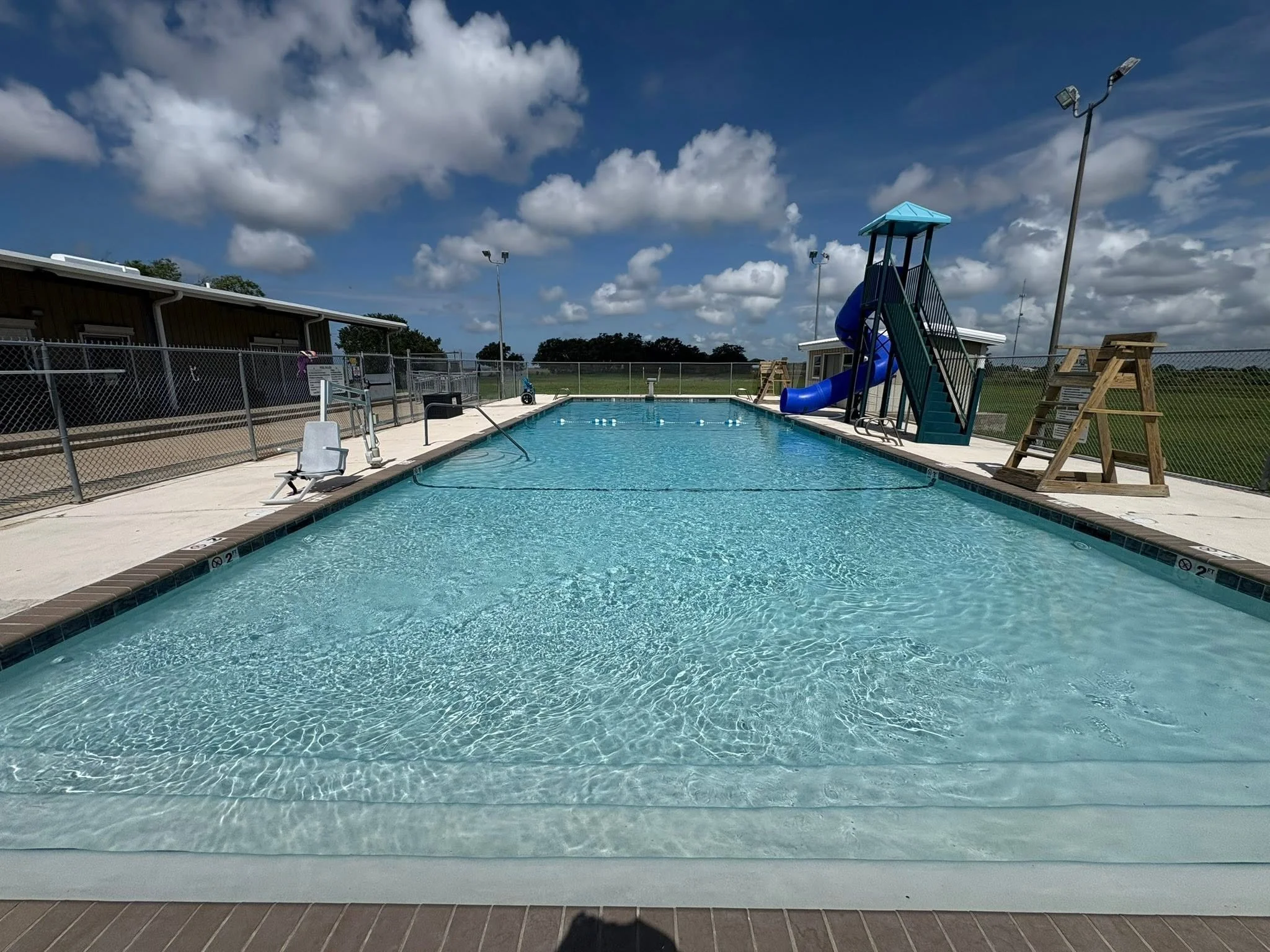 Empty outdoor swimming pool with a blue water slide, surrounded by a chain-link fence and a building, under a partly cloudy sky.