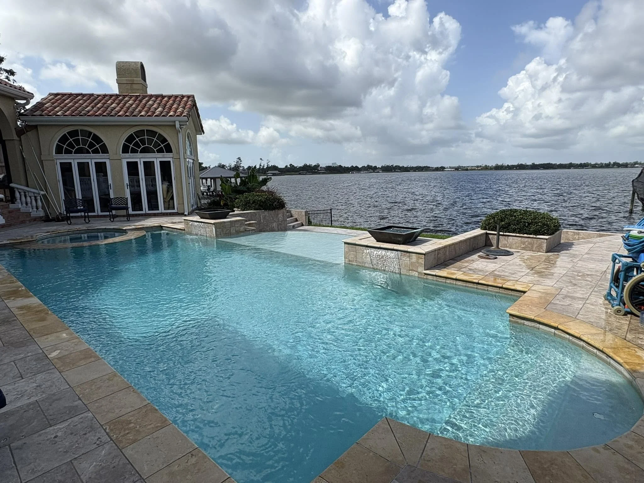 A backyard with a swimming pool overlooking a large body of water and houses in the distance under a partly cloudy sky.