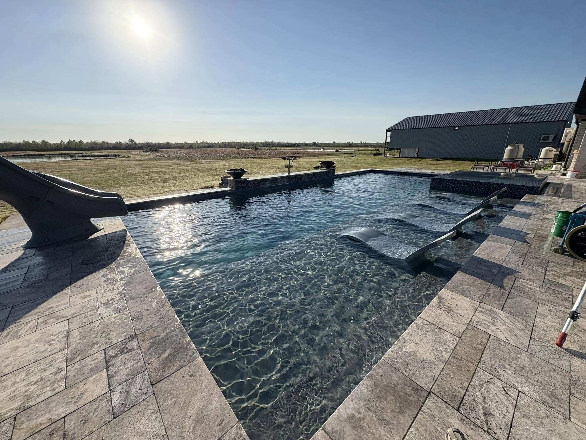 A backyard scene showing a clean swimming pool with water slides, a patio area with outdoor furniture, an open field with tall grass, trees, and a barn-like structure in the background, under a clear blue sky and bright sunlight.