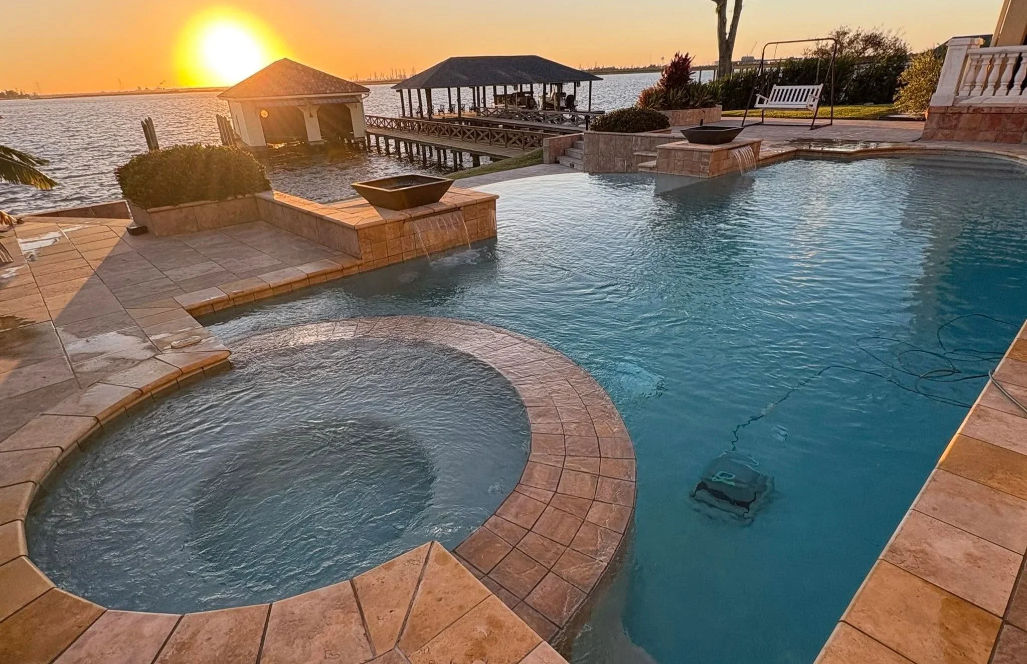 A scene at sunset showing a backyard with a pool and hot tub overlooking a body of water. There are structures including a gazebo and a dock with a gazebo, with chairs inside. The area has landscaping with trees and bushes, and a swing seat is present near the house.