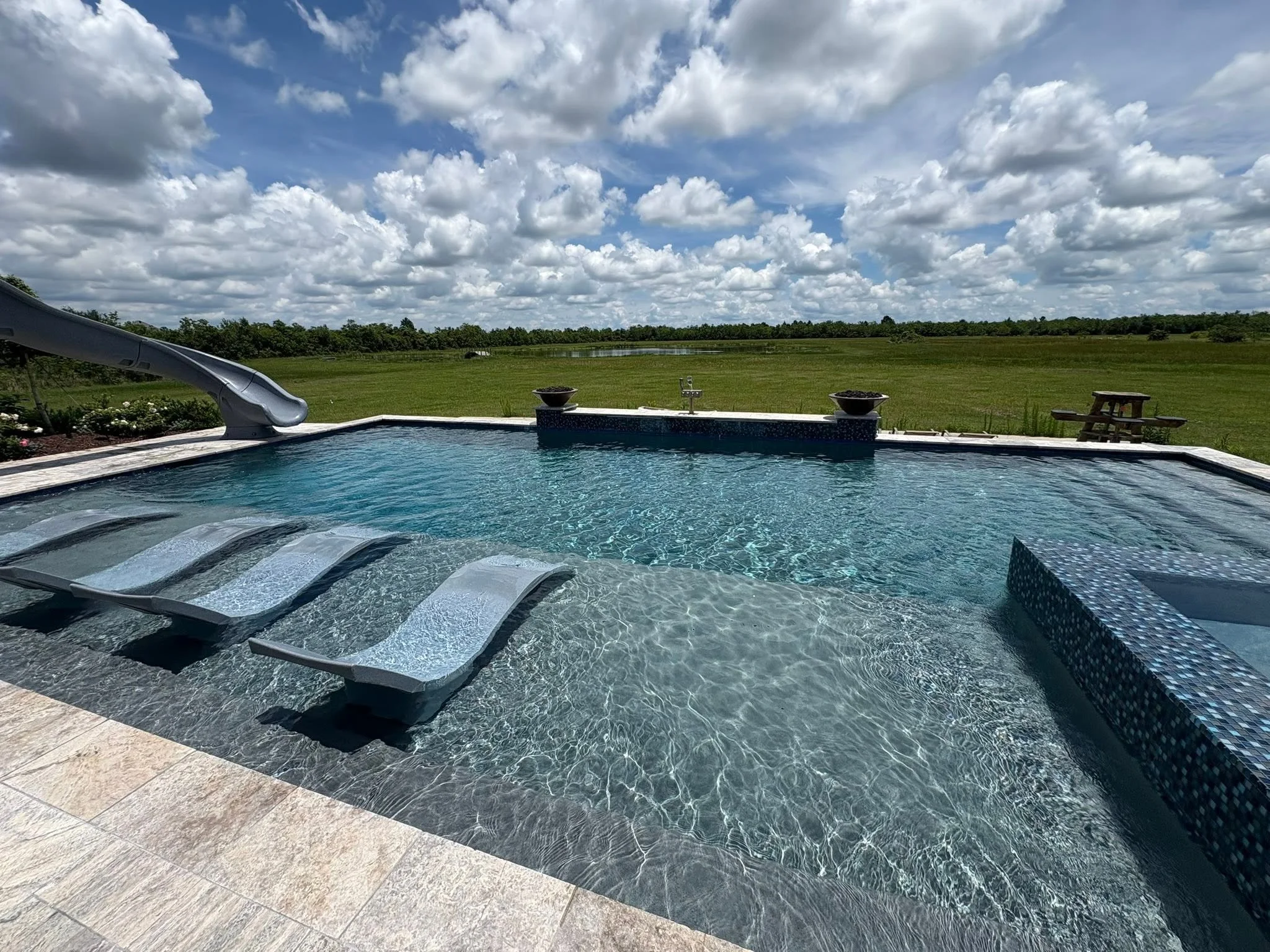 An outdoor swimming pool with a slide in the corner, surrounded by a stone deck. Beyond the pool, there is a large grassy field with a small pond and a line of trees in the distance under a partly cloudy sky.