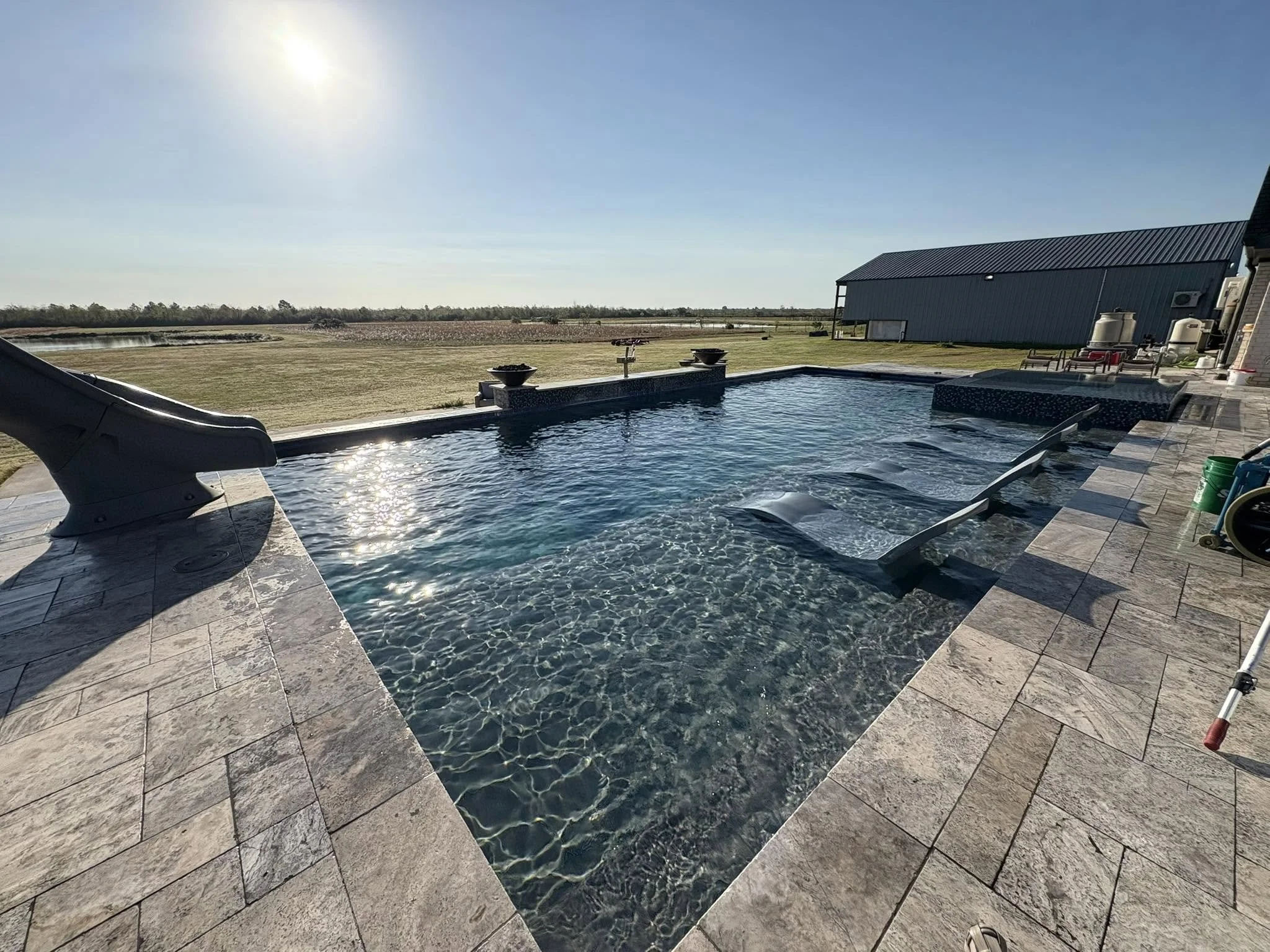 Outdoor backyard with a swimming pool, lounge chairs, and a barn-like structure, with a vast open field and blue sky in the background.