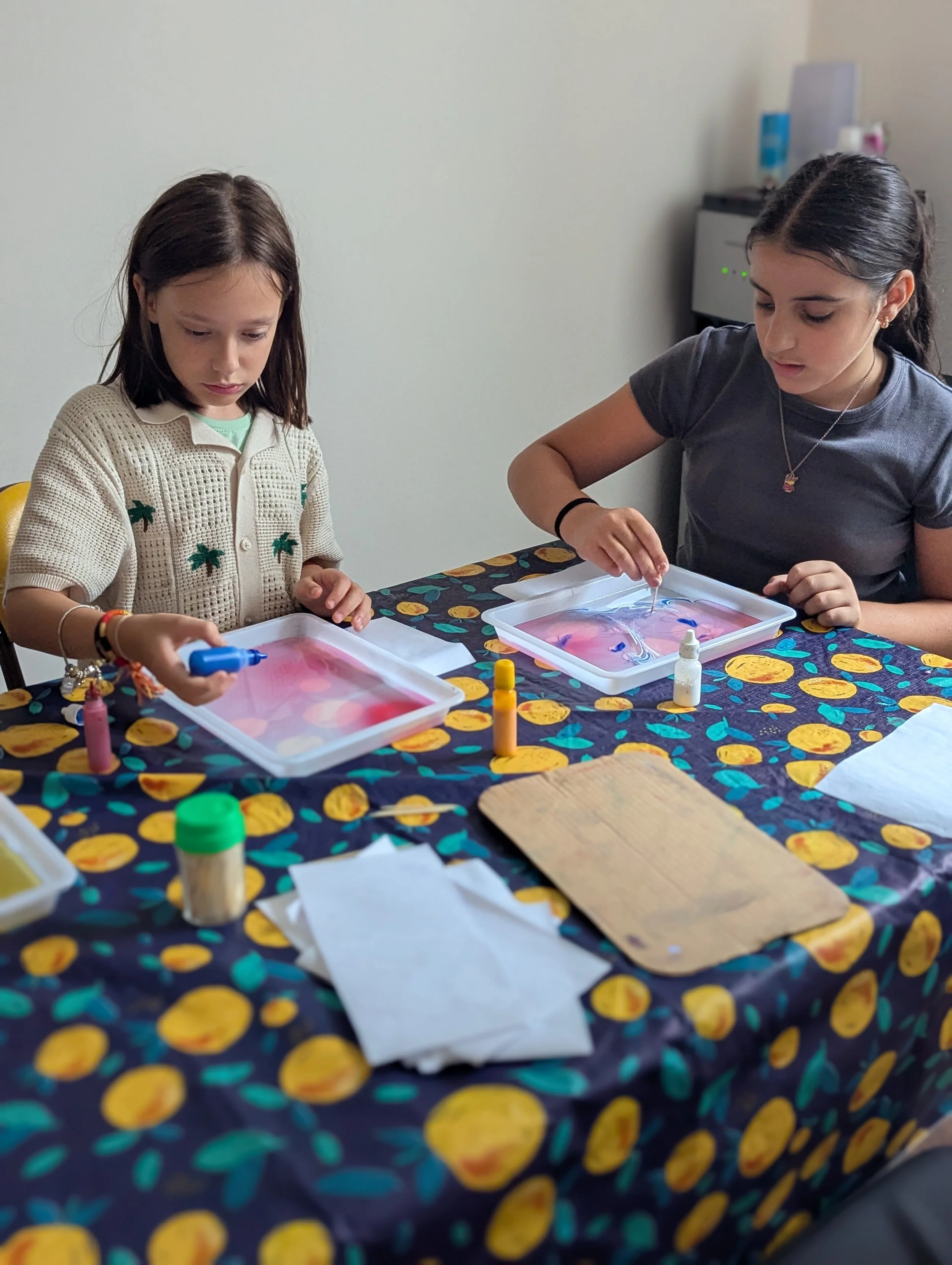 Two young girls engaged in a marbling activity at a table with colorful tablecloth and supplies during MASKIT, Ashreynu's experiential Hebrew School program.
