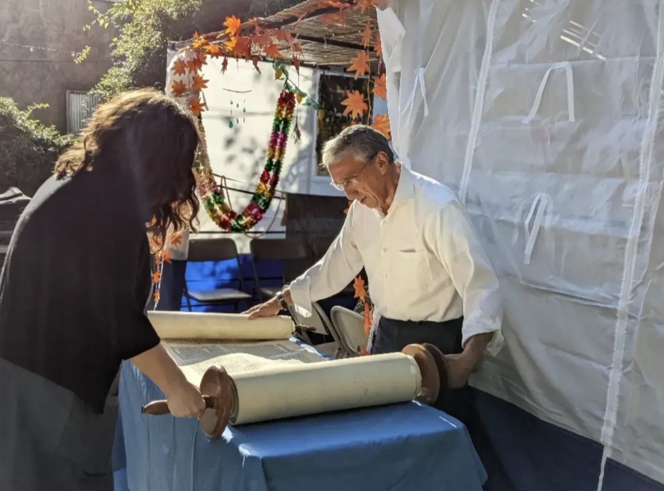 A man and a woman are standing at a table with a Torah scroll during a Sukkot celebration, with autumn-themed decorations and a floral wreath hanging in the background.