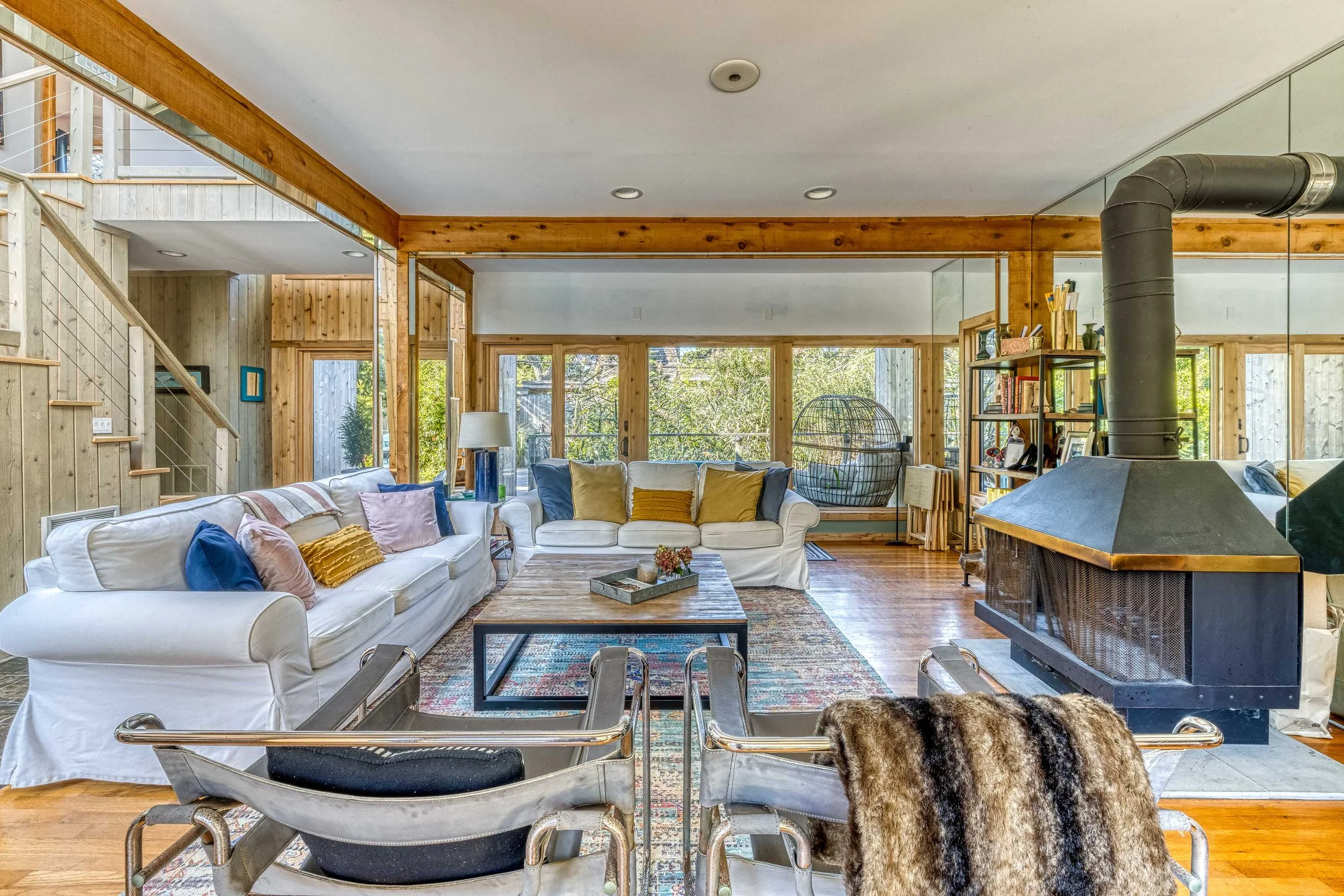 Living room with wooden accents, white sofas with colorful pillows, large windows showing trees outside, a fireplace, a bookshelf, and a glass hanging chair.