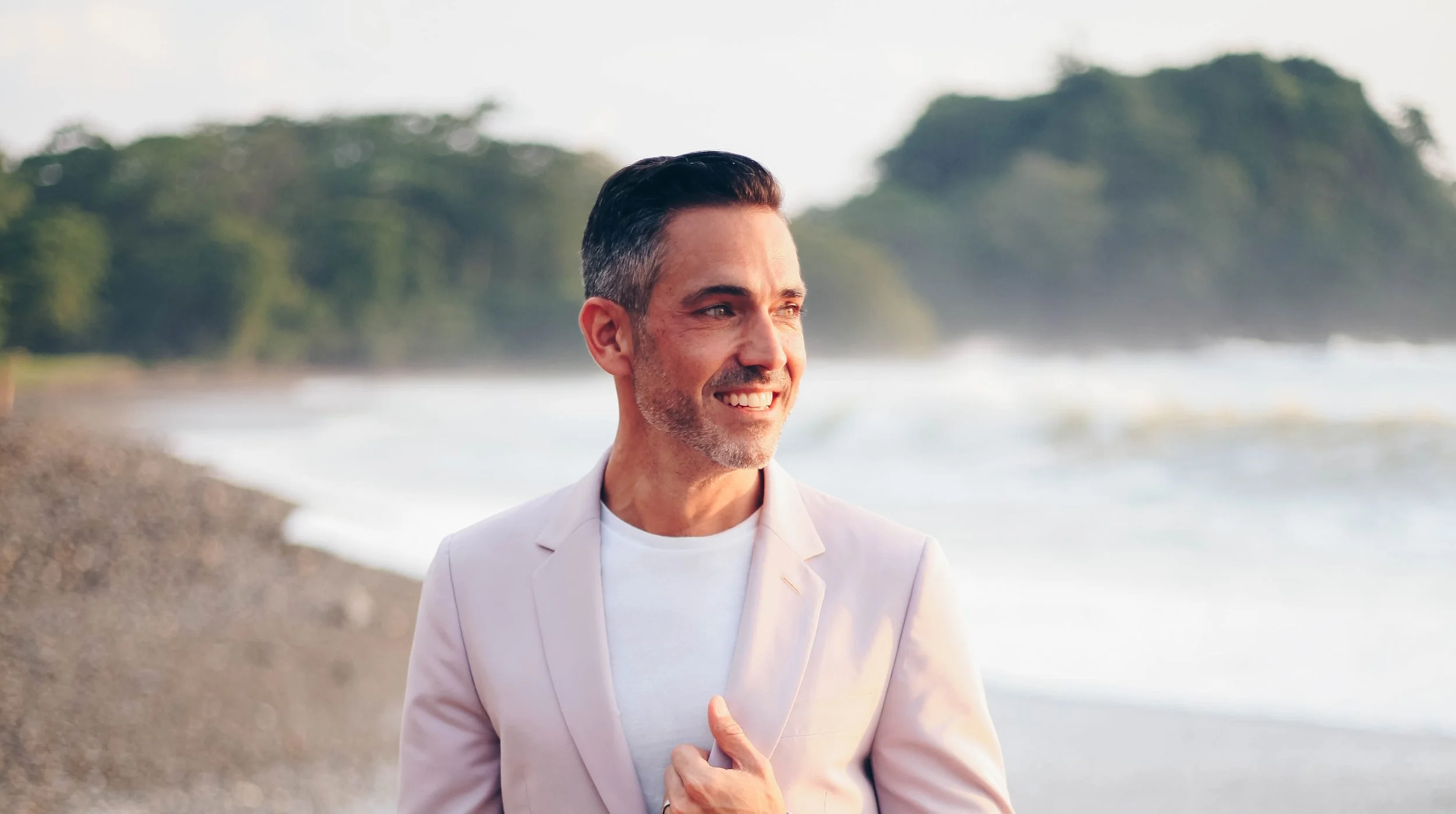 A middle-aged man with dark hair and a gray beard, smiling, near a beach with trees in the background, dressed in a light pink blazer and white shirt.