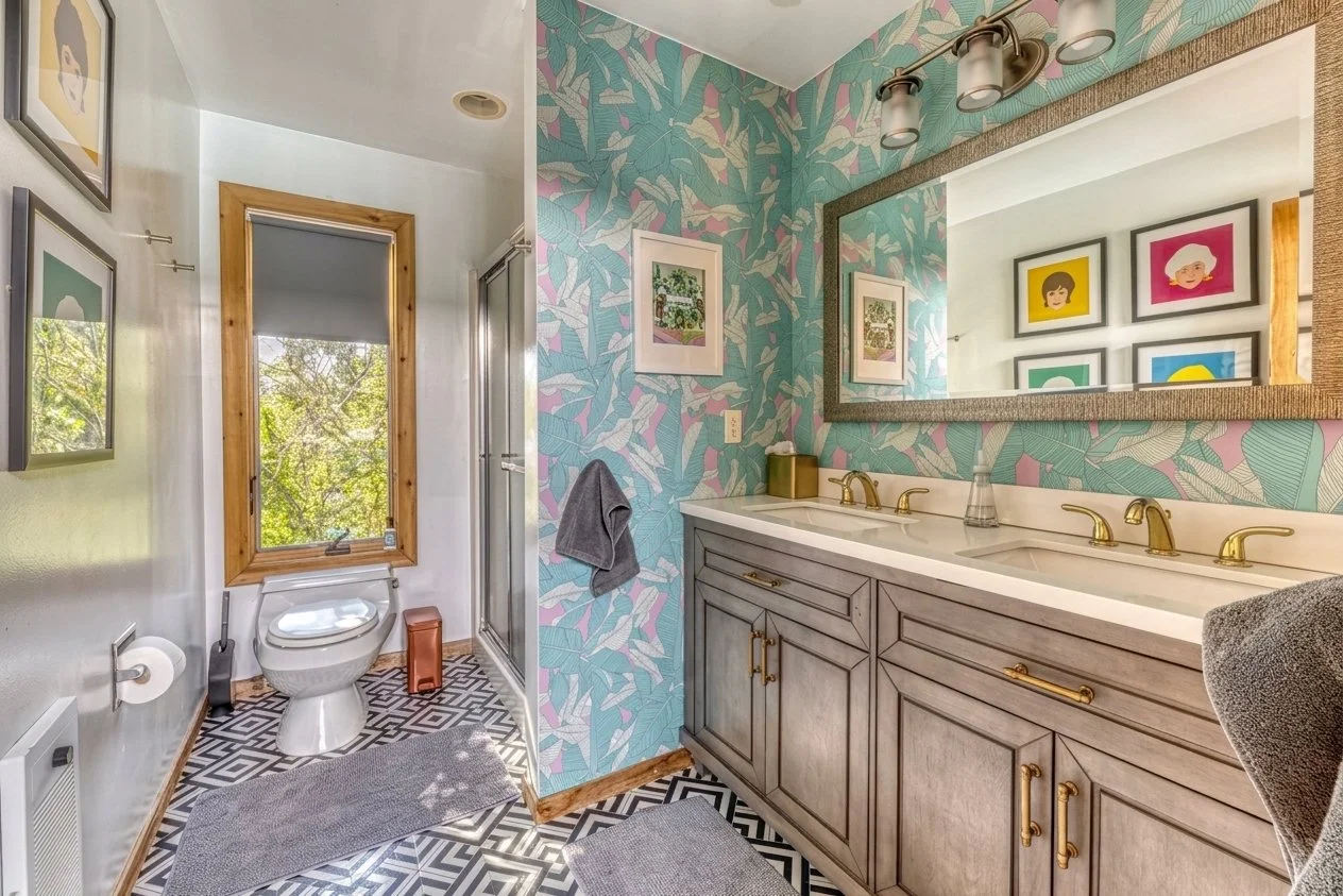 A bathroom featuring a wooden-framed window, a toilet, and a shower with glass door. The walls have colorful, tropical leaf patterned wallpaper. There are framed artworks and a large mirror above a double vanity with gold fixtures. The floor has black and white geometric tiles, and gray rugs are placed in front of the vanity and toilet.