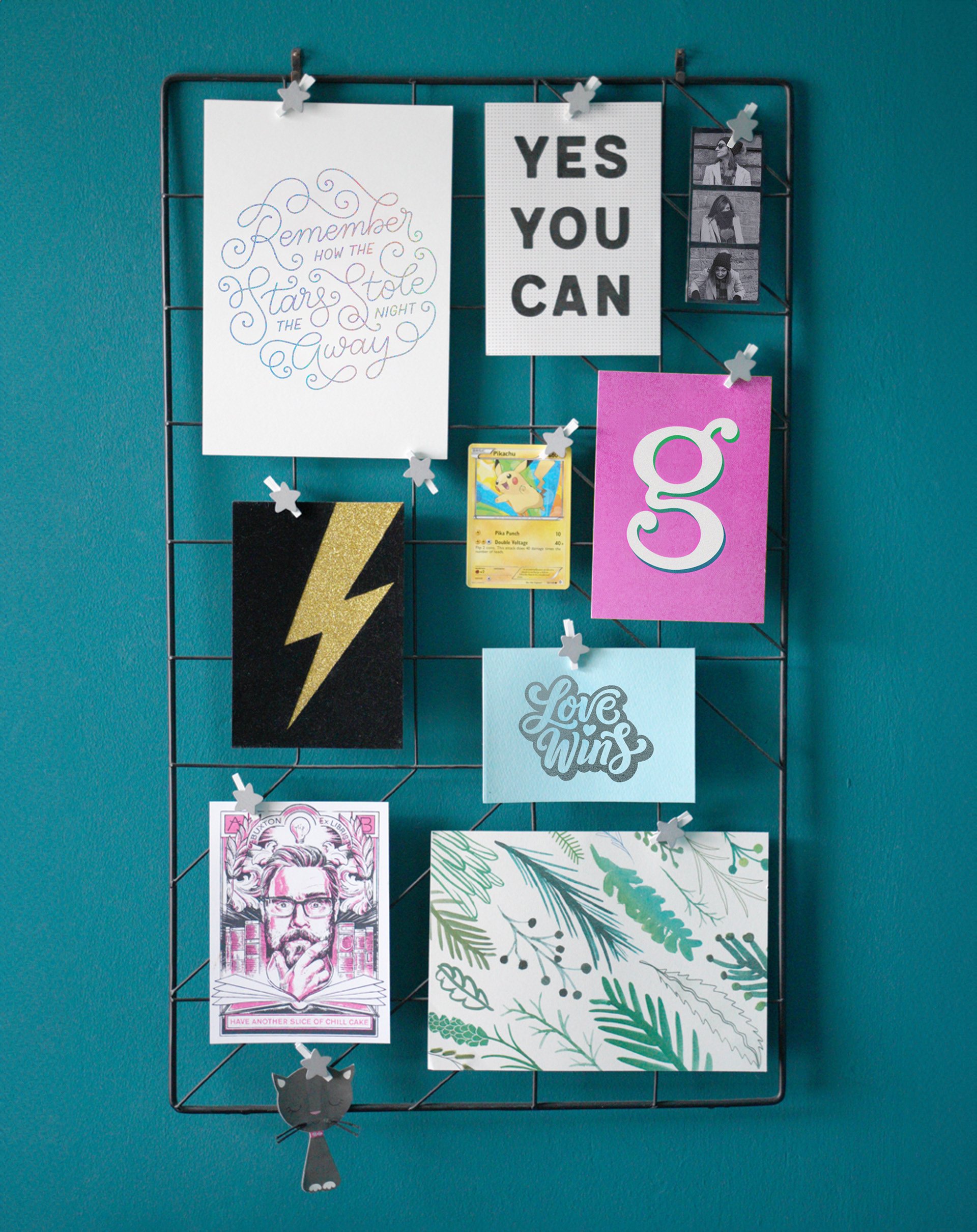 A black wire grid hanging on a teal wall displays various decorative items, including printed motivational quotes, black and white photos, a Pokémon card, a patterned postcard, and a black cat-shaped paper clip. The quotes include 'Remember how the stars stole the night away,' 'YES YOU CAN,' and 'Love Wins,' along with a colorful illustrated cartoon of Pikachu.
